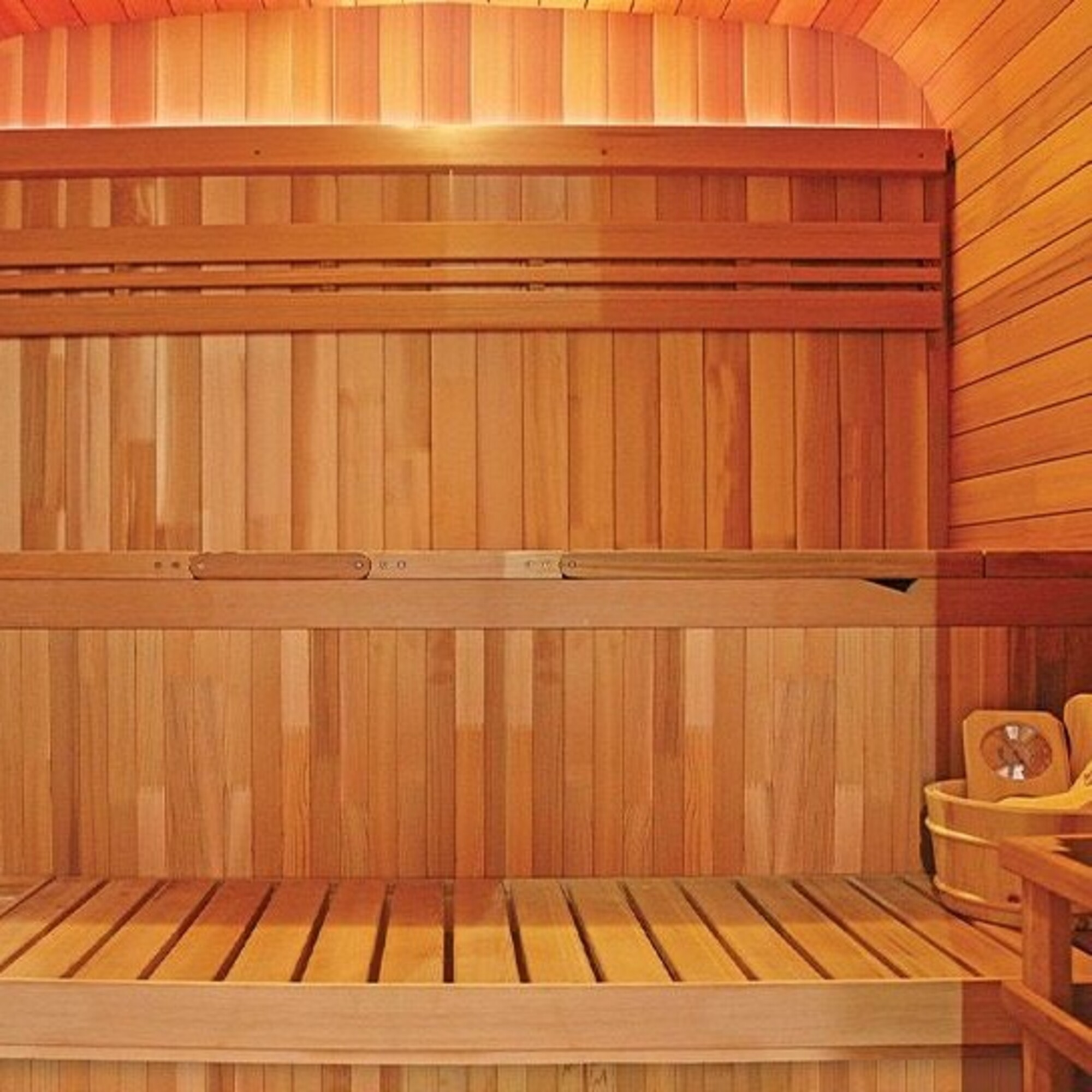 Interior de sauna de madera. Paneles verticales de madera, bancos y techo arqueado. Se ven un cubo y un temporizador.