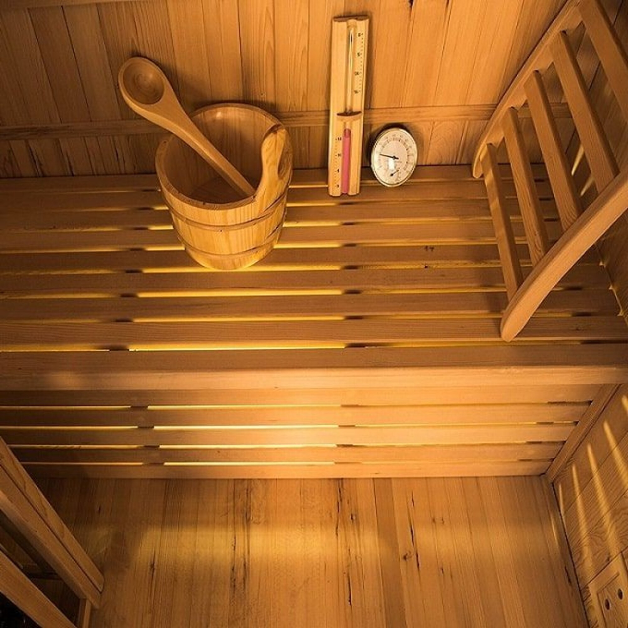 Interior de sauna de madera con un cubo, cucharón, termómetro y silla. La escena está iluminada con luz cálida.