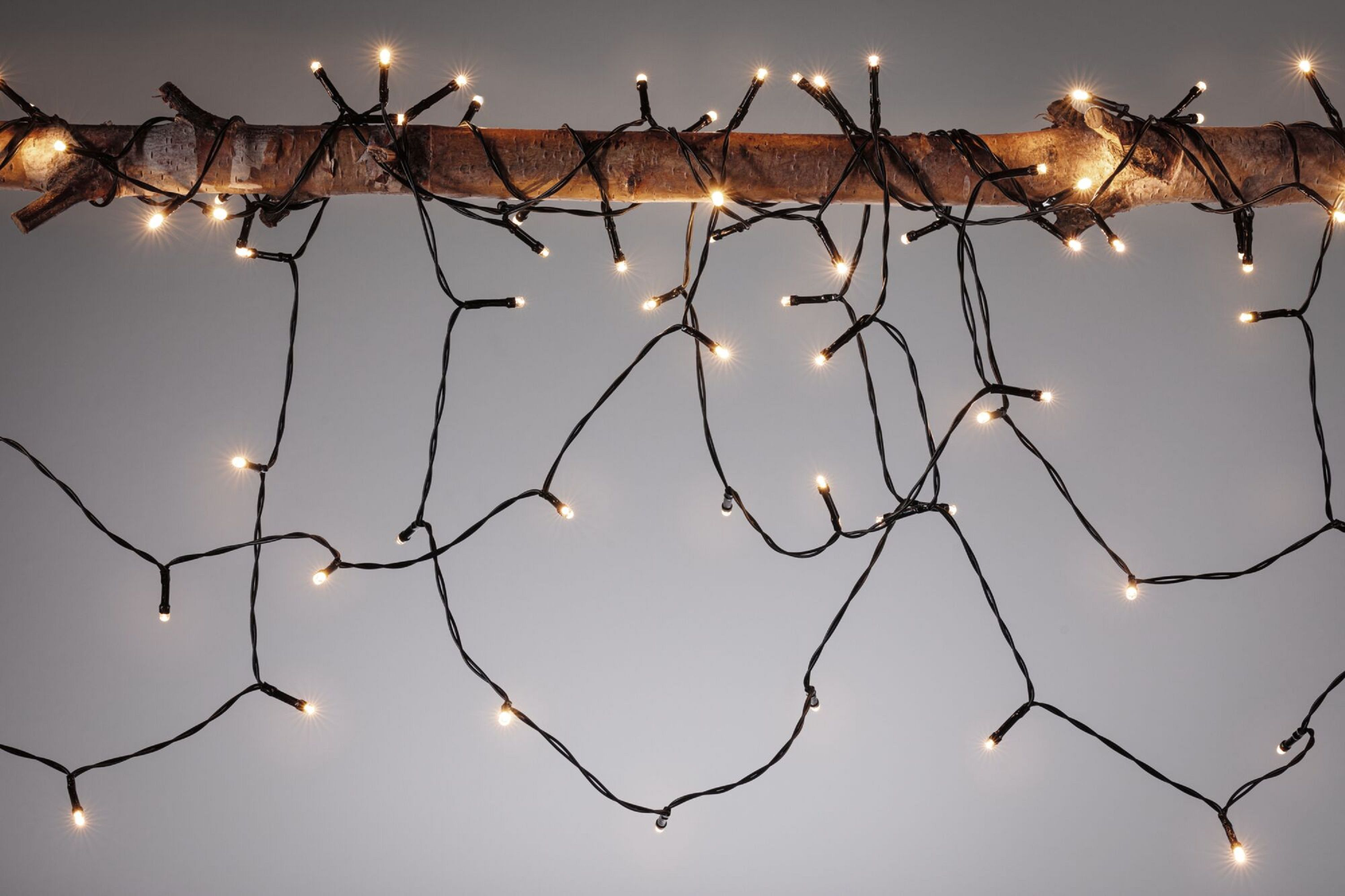Ein Ast mit warmweißen Lichterketten. Schwarze Drähte fallen herab und erzeugen ein festliches Leuchten vor grauem Hintergrund.