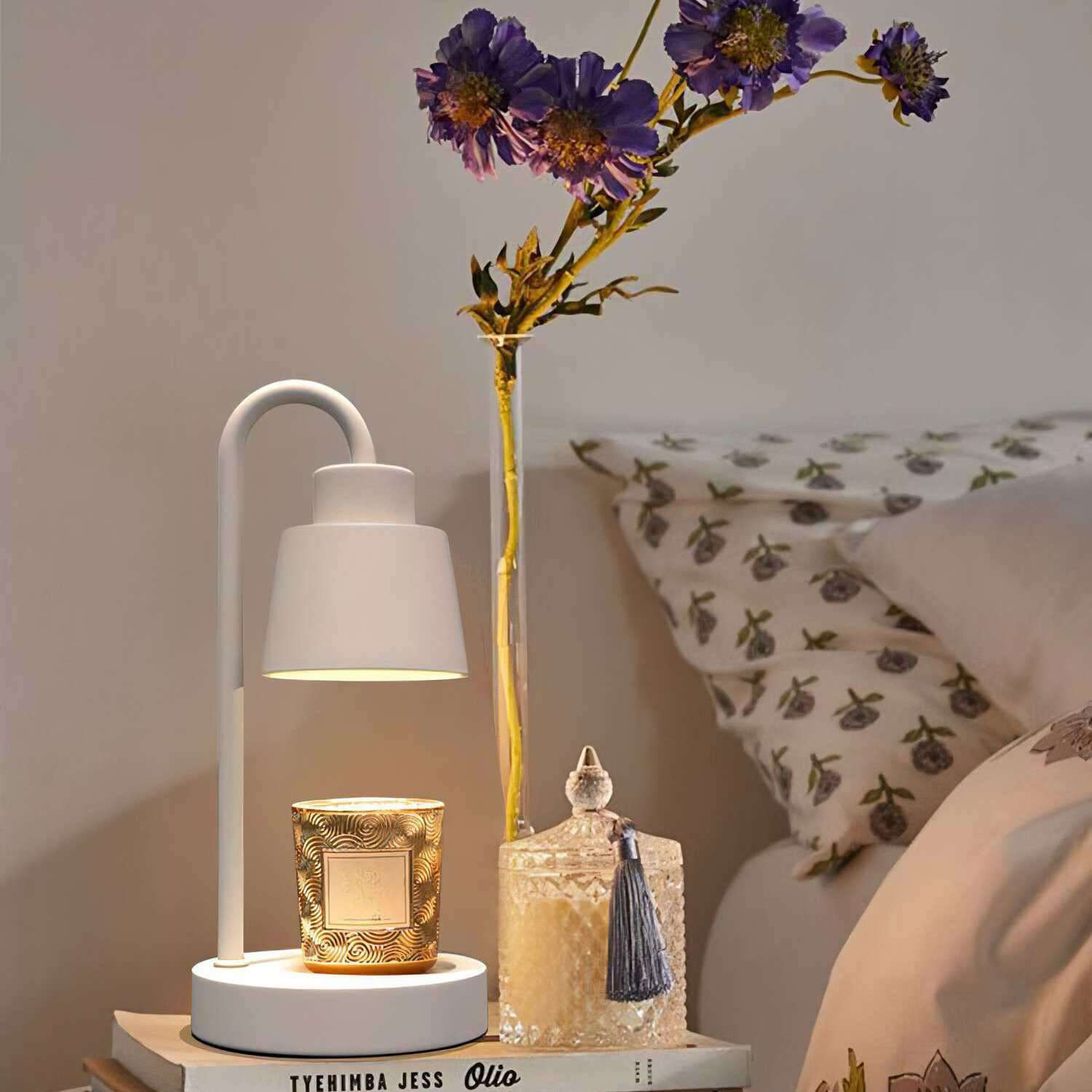 Eine weiße Lampe mit einer Kerze und Blumen. Der Hintergrund ist ein Bett mit Kissen. Die Lampe steht auf einem Buch.