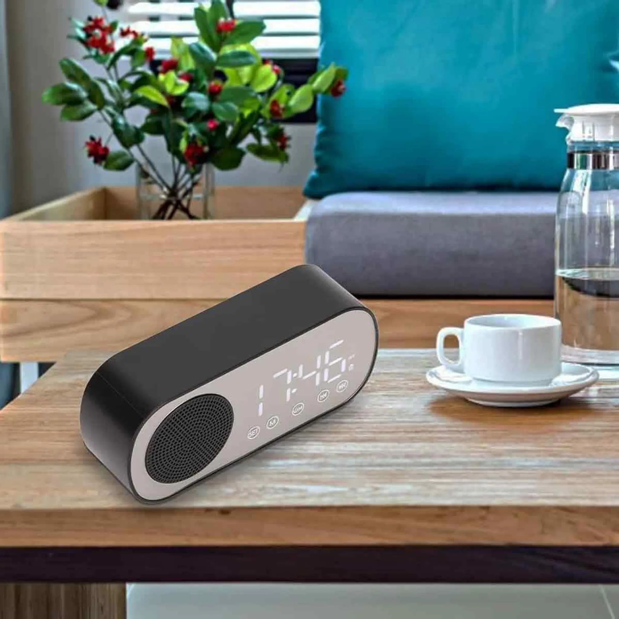 Schwarzes Radiowecker auf einem Holztisch, neben einer weißen Tasse und Untertasse. Hintergrund umfasst Pflanzen und ein blaues Kissen.