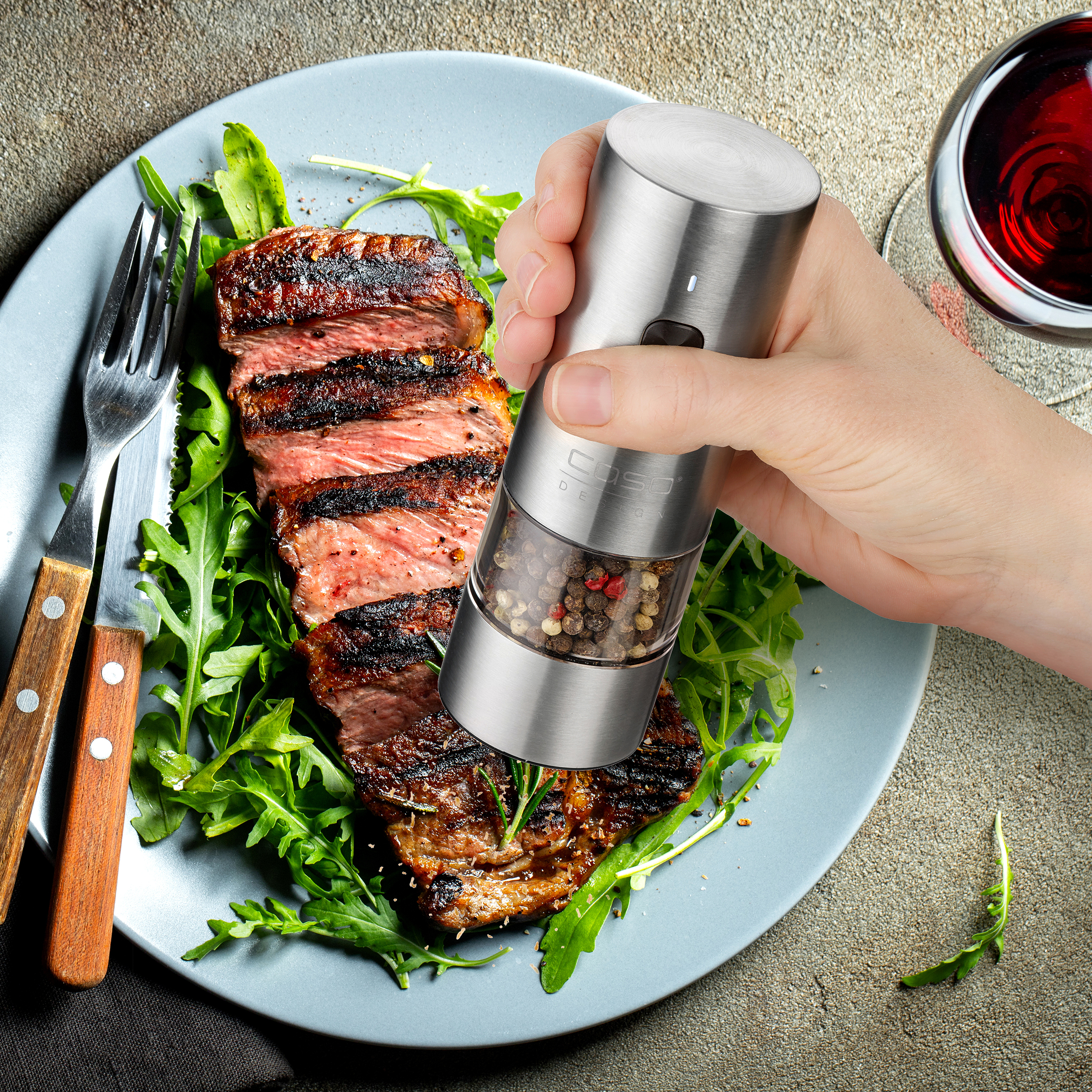 Eine Hand mahlt Pfeffer auf ein gegrilltes Steak. Das Steak liegt auf einem Teller mit Grünzeug, Gabel und Messer.