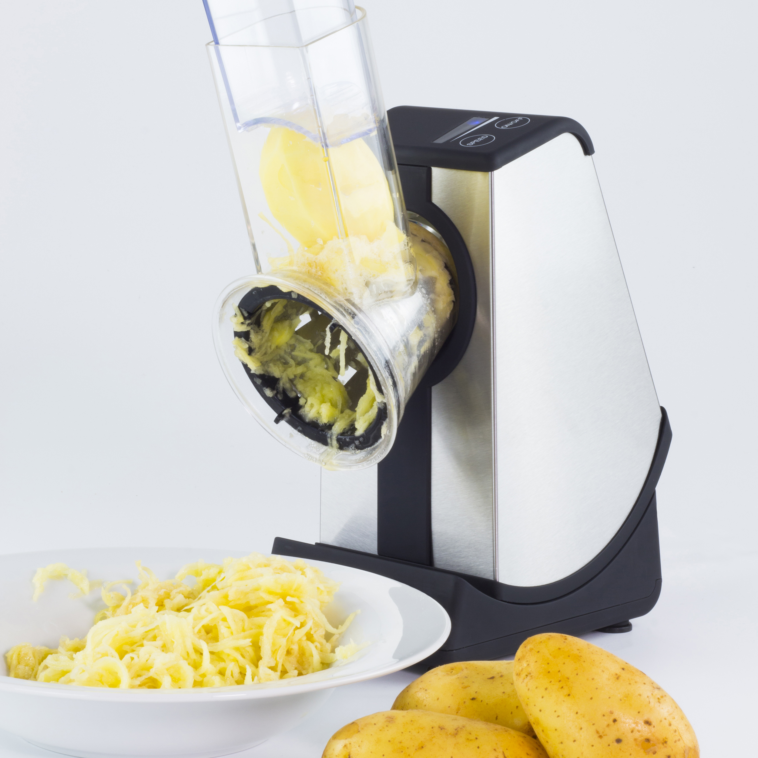 Ein Kartoffelschneider raspelt Kartoffeln in eine weiße Schüssel. Ganze Kartoffeln liegen im Hintergrund auf dem Tisch.