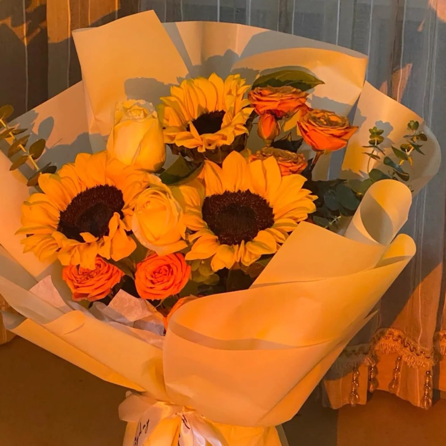 Ein Blumenstrauß mit Sonnenblumen, Rosen und Grünzeug, eingewickelt in gelbes Papier, gebunden mit einem weißen Band, vor einem Vorhanghintergrund.