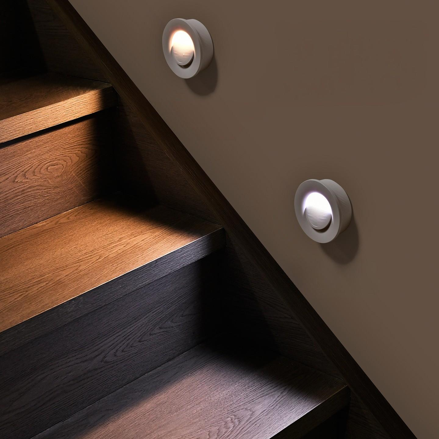 Holztreppe mit zwei Wandleuchten. Die Stufen sind braun und dunkel. Die Leuchten sind weiß, kreisförmig und strahlen Licht aus.