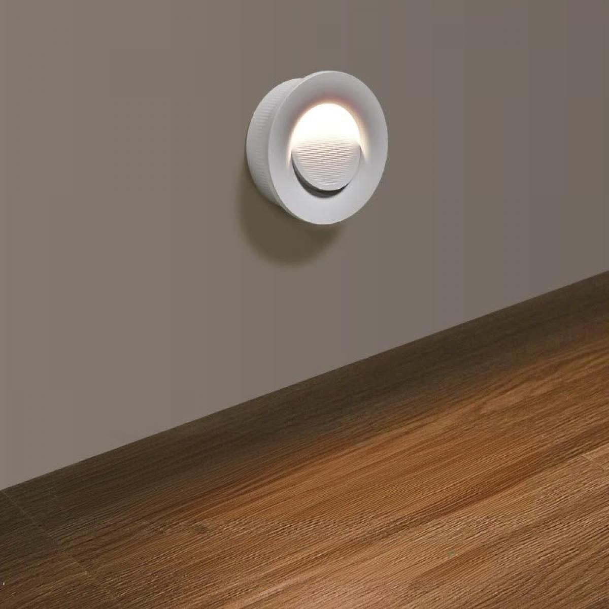 Eine weiße, kreisförmige Wandleuchte beleuchtet einen Raum. Das Licht ist an einer grauen Wand angebracht, mit Holzboden im Vordergrund.