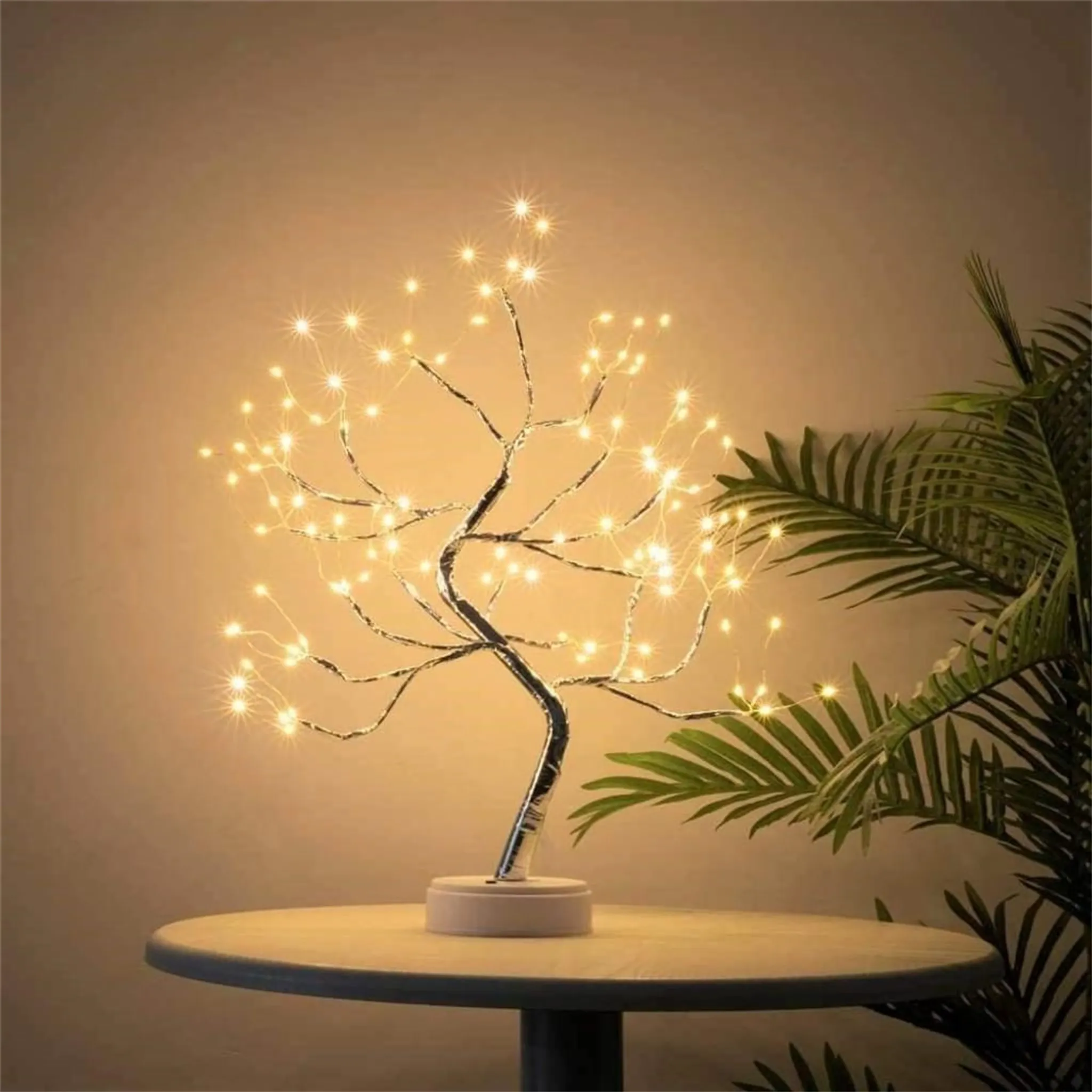Ein dekorativer Baum mit Lichtern steht auf einem Tisch. Eine Pflanze befindet sich neben dem Baum, eine Wand im Hintergrund.