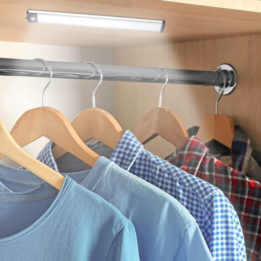 In einem Schrank beleuchtet ein Licht Kleidung auf Kleiderbügeln. Hemden und T-Shirts hängen an einer Metallstange. Holzaufhänger sind sichtbar.