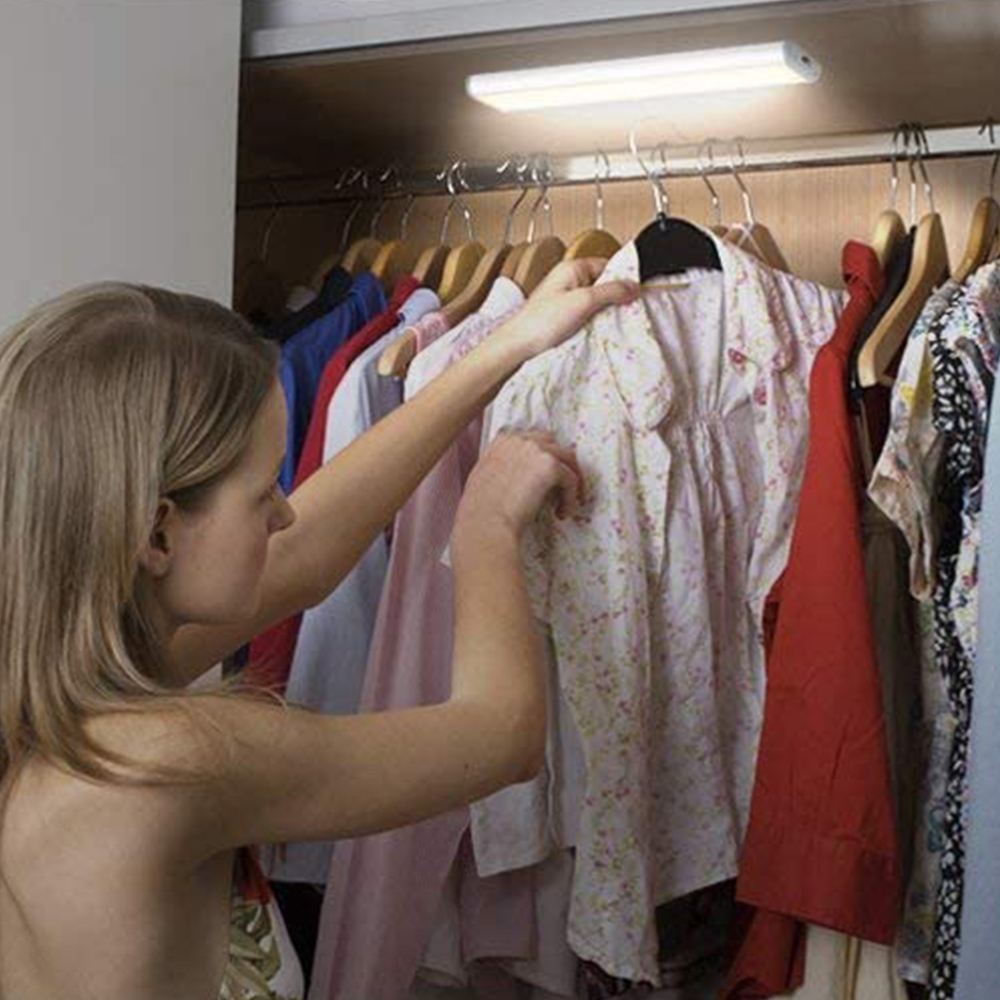 Eine Frau wählt in einem Schrank ein geblümtes Hemd aus. Kleidung hängt an Kleiderbügeln. Über der Kleidung befindet sich ein Licht.