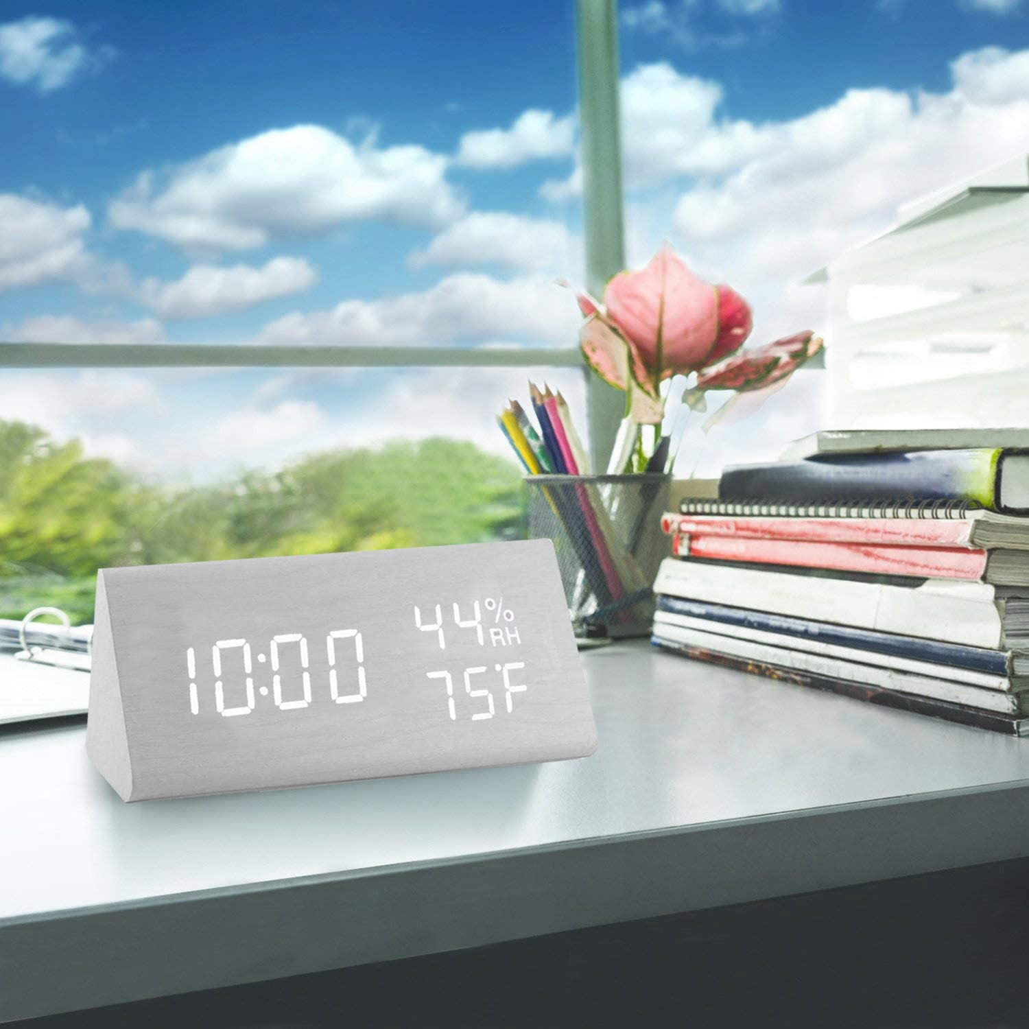 Ein Schreibtisch mit digitaler Uhr, Blumen und Bleistiften. Ein Fenster zeigt blauen Himmel mit Wolken und grüner Landschaft.
