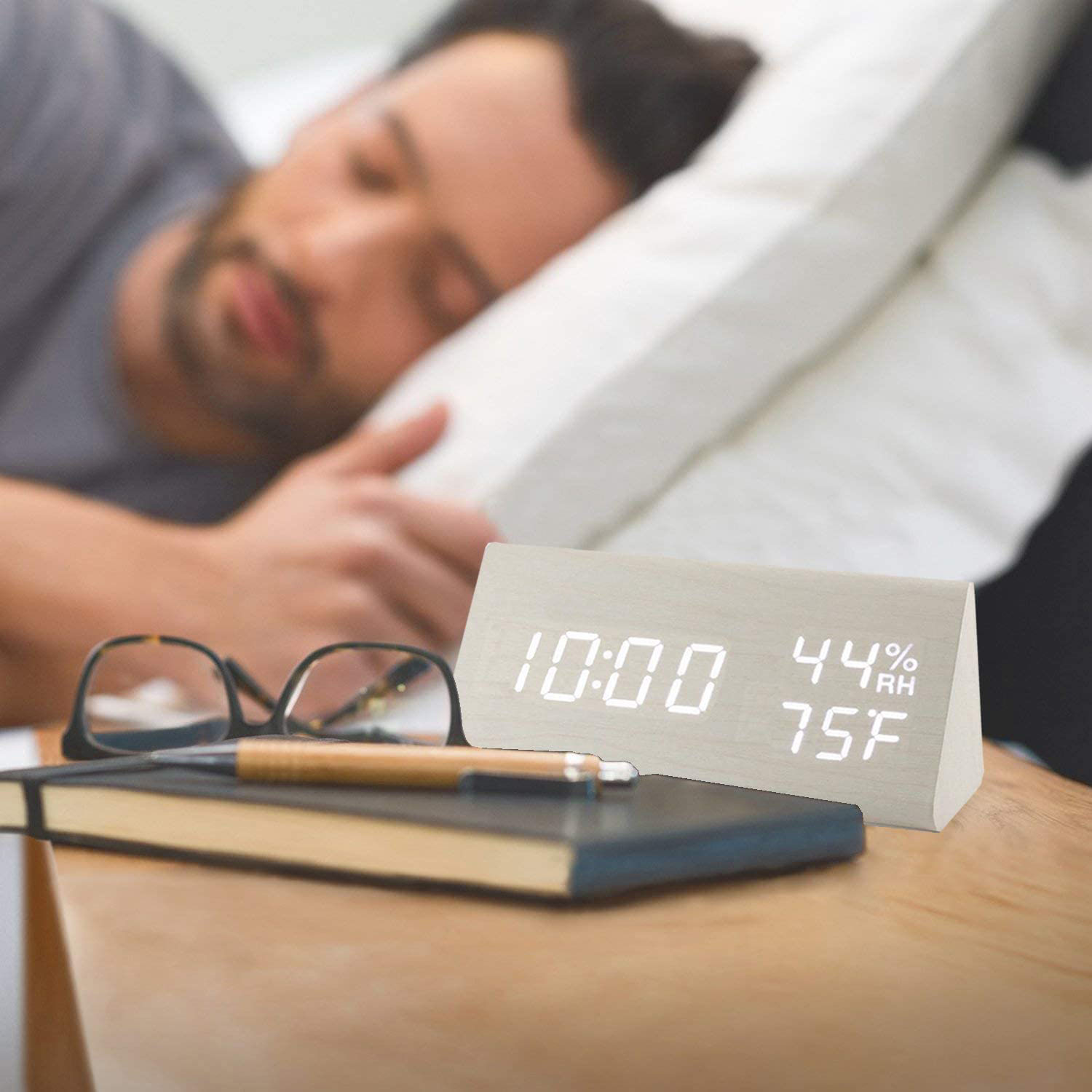 Mann schläft im Bett. Daneben zeigt eine Holzuhr Zeit, Luftfeuchtigkeit und Temperatur an. Brille, Stift und Notizbuch liegen auf dem Tisch.