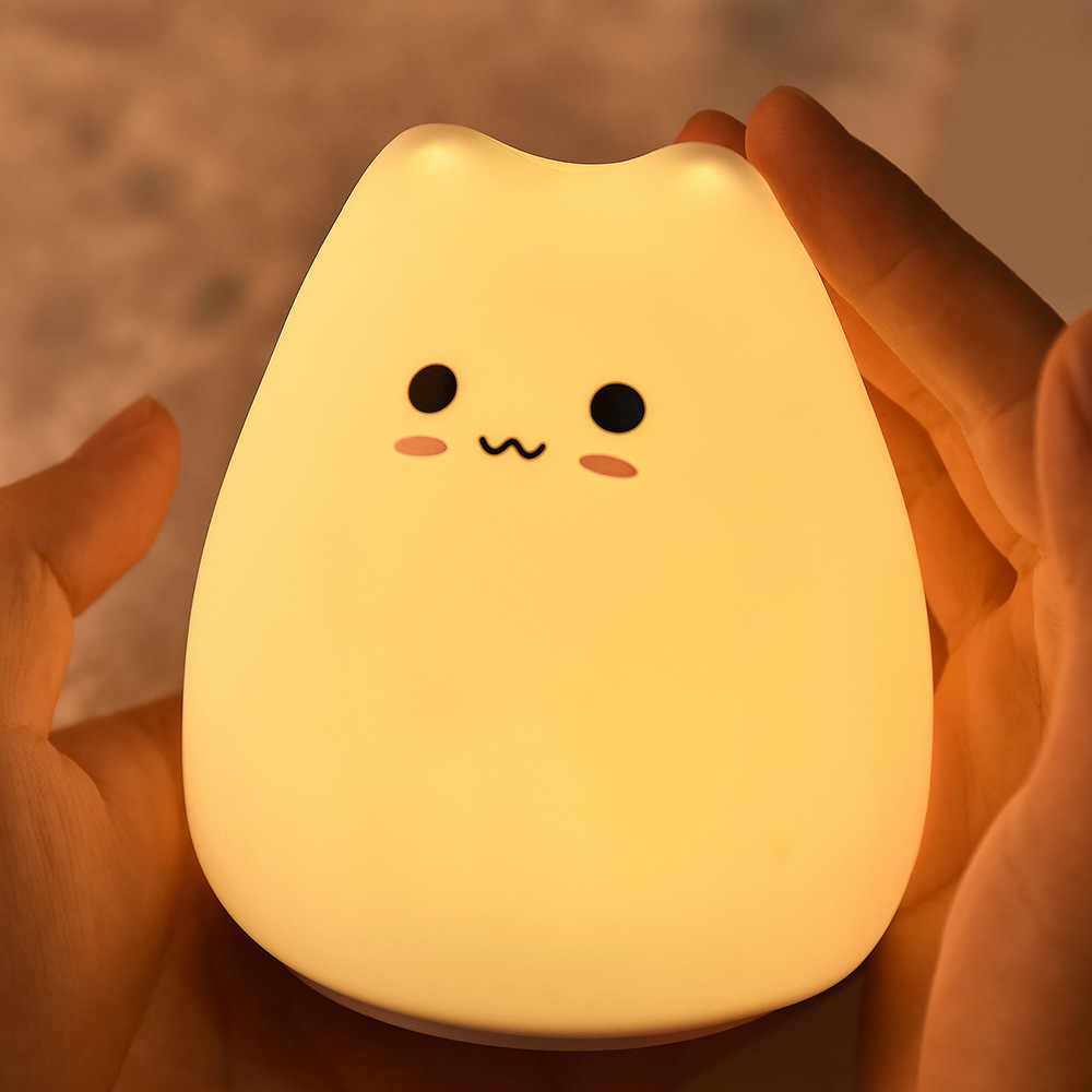 Ein leuchtendes, katzenförmiges Nachtlicht, das von zwei Händen gehalten wird. Das Licht ist gelb mit einem süßen Gesicht. Der Hintergrund ist verschwommen und warm.