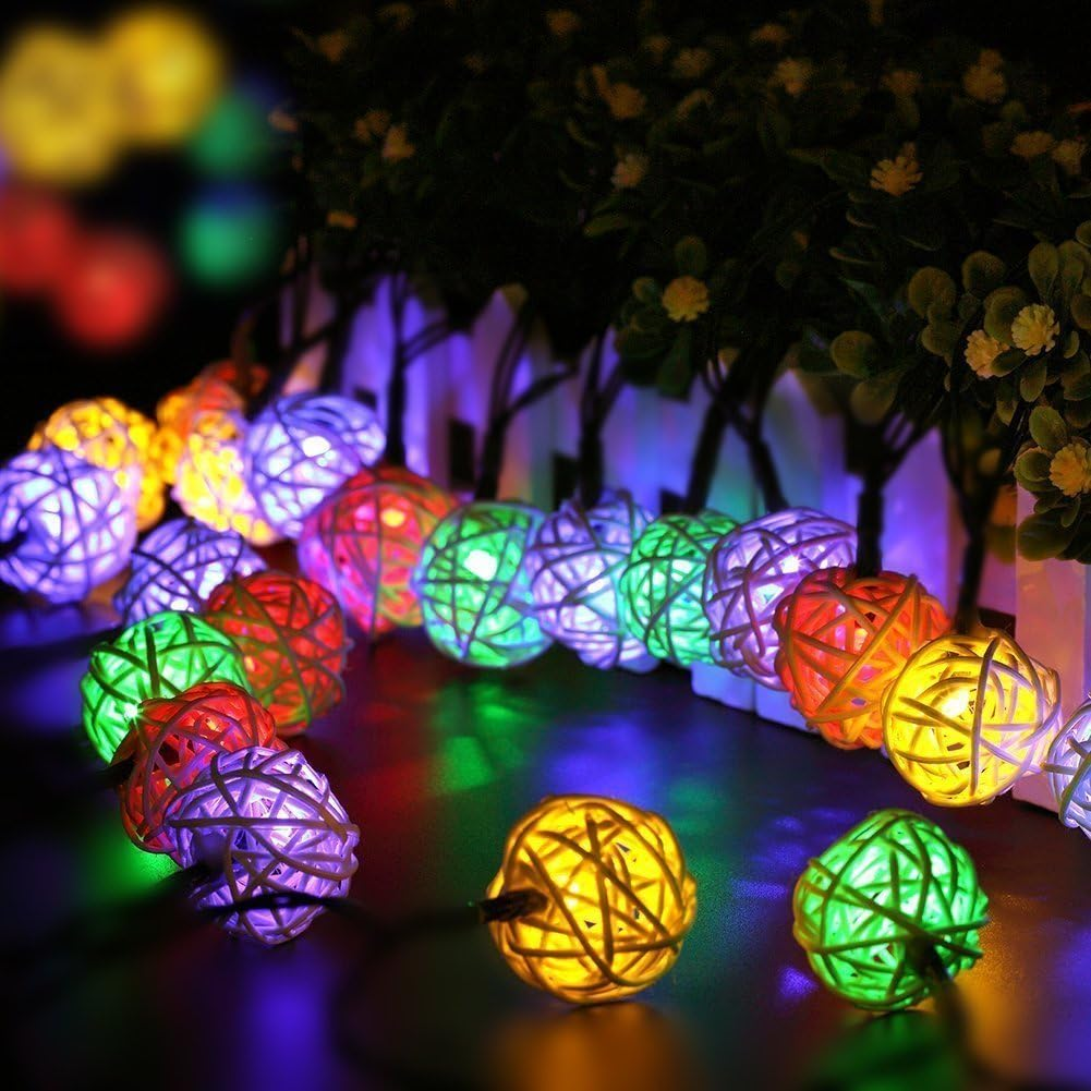 Lichterketten aus farbenfrohen gewebten Kugeln. Grüne, rote, gelbe und violette Lichter im Vordergrund. Grüne Pflanzen und ein weißer Zaun im Hintergrund.