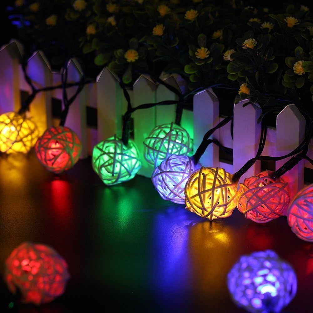 Lichterketten mit bunten gewebten Kugeln vor einem weißen Zaun, dahinter Grün. Lichter spiegeln sich auf der Oberfläche.