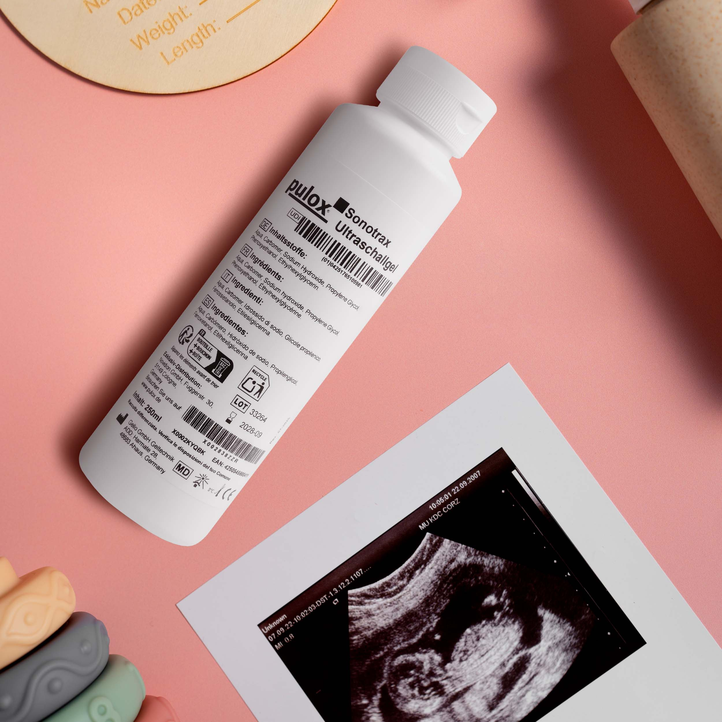 Eine Flasche Ultraschallgel, ein Ultraschallbild und Babyspielzeug auf rosa Hintergrund.