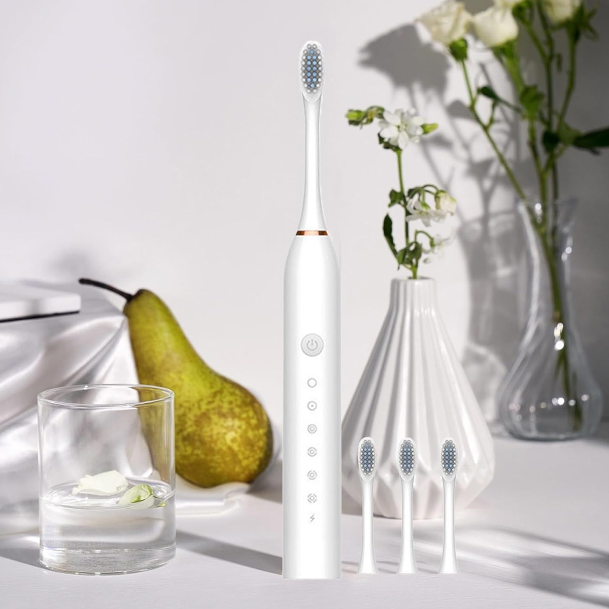 Eine weiße elektrische Zahnbürste steht mit drei Bürstenköpfen. Eine Birne, ein Glas Wasser und Blumen in Vasen sind ebenfalls vorhanden.