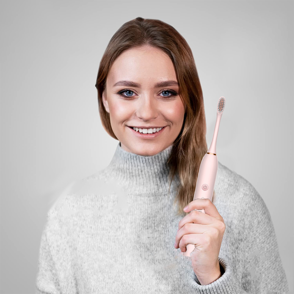 Eine lächelnde Frau hält eine rosa elektrische Zahnbürste. Sie trägt einen grauen Pullover vor grauem Hintergrund.