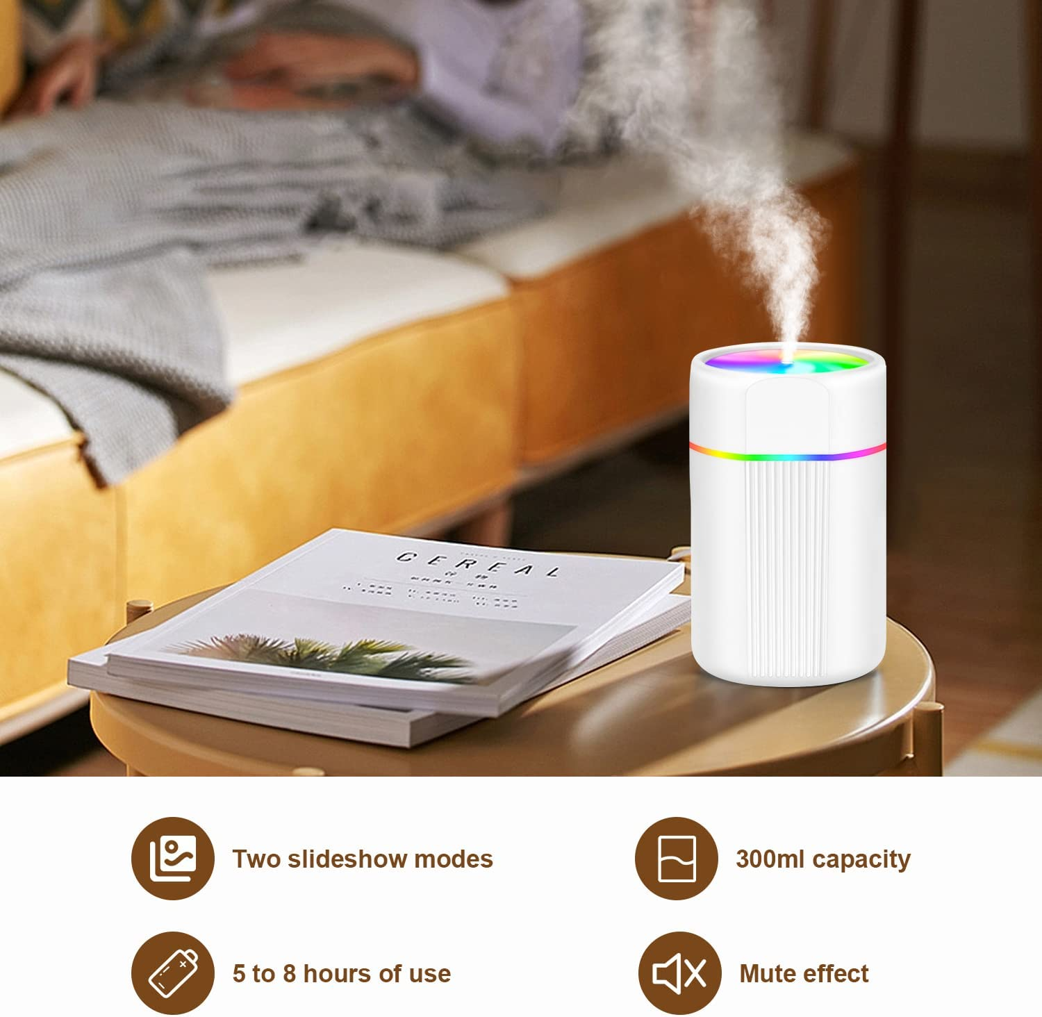 Ein weißer Luftbefeuchter mit Regenbogenlichtern stößt Dampf aus. Bücher und ein Tisch befinden sich im Vordergrund. Im Hintergrund ist ein Bett.