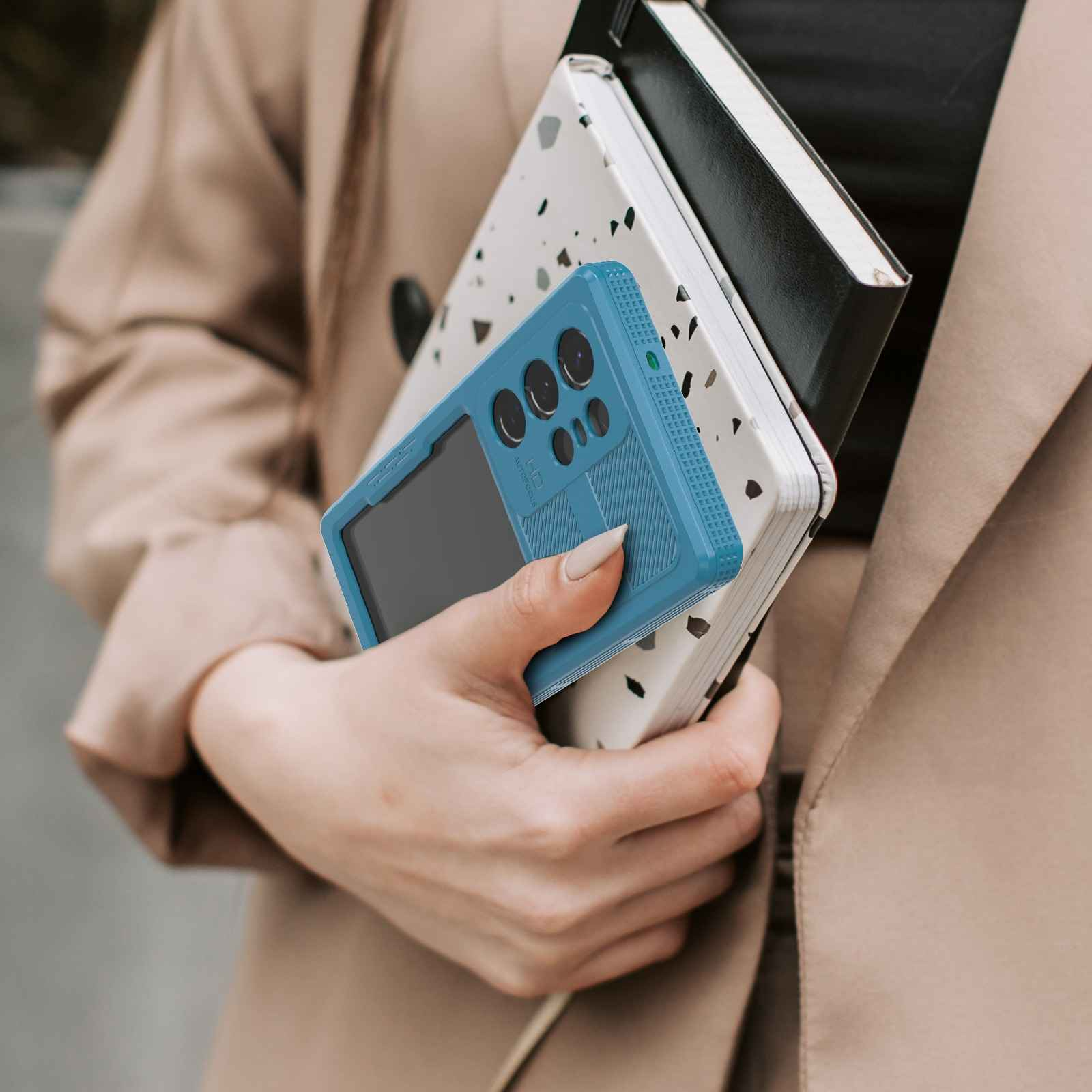 Eine Person hält eine blaue Handyhülle und zwei Notizbücher. Die Person trägt einen beigefarbenen Mantel. Die Notizbücher sind weiß und schwarz.