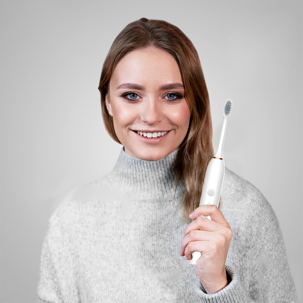 Eine lächelnde Frau hält eine weiße elektrische Zahnbürste. Sie trägt einen grauen Pullover vor grauem Hintergrund.