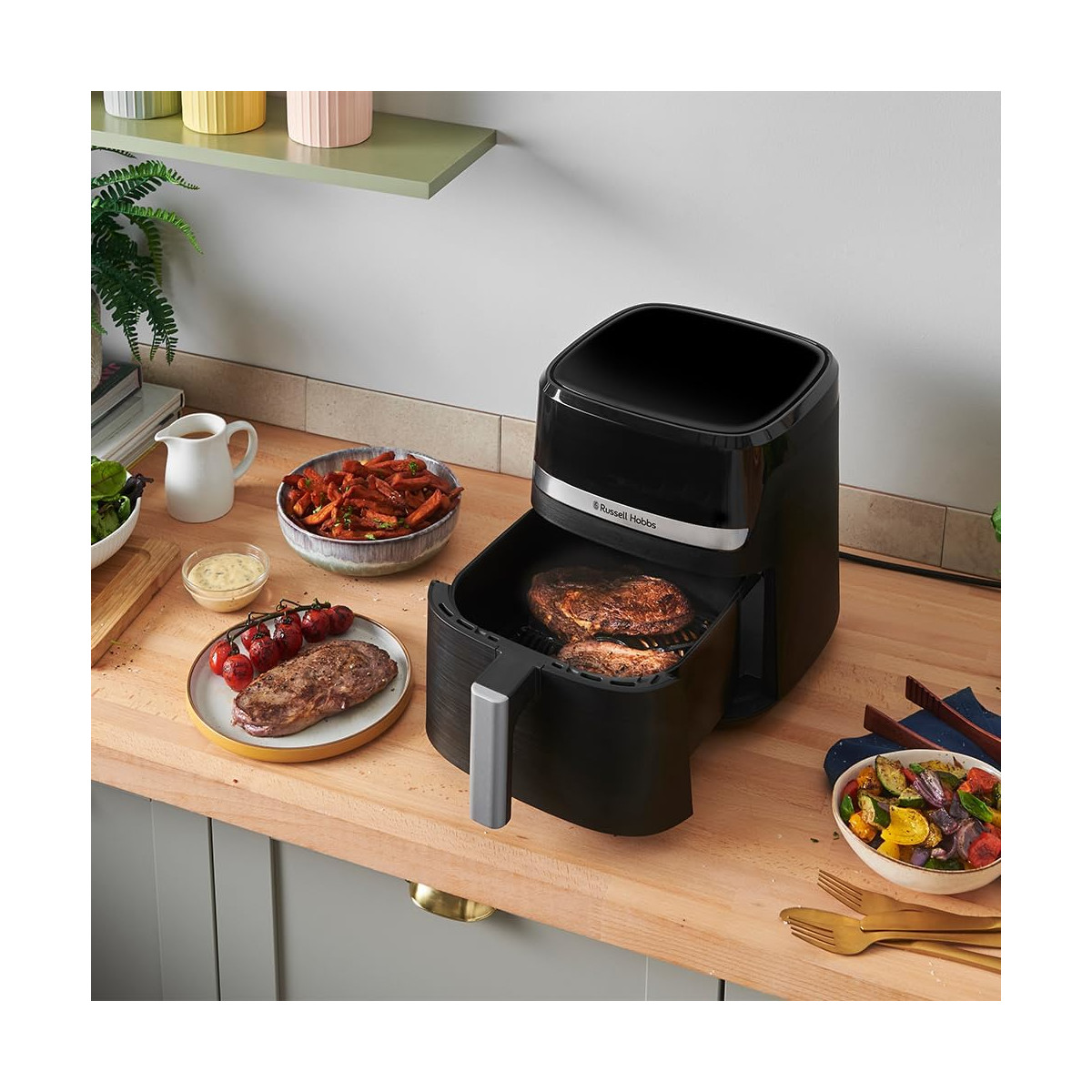 Ein schwarzer Airfryer mit gebratenen Steaks, umgeben von vorbereiteten Speisen. Gemüse, Saucen und Utensilien liegen auf einer Holzplatte.