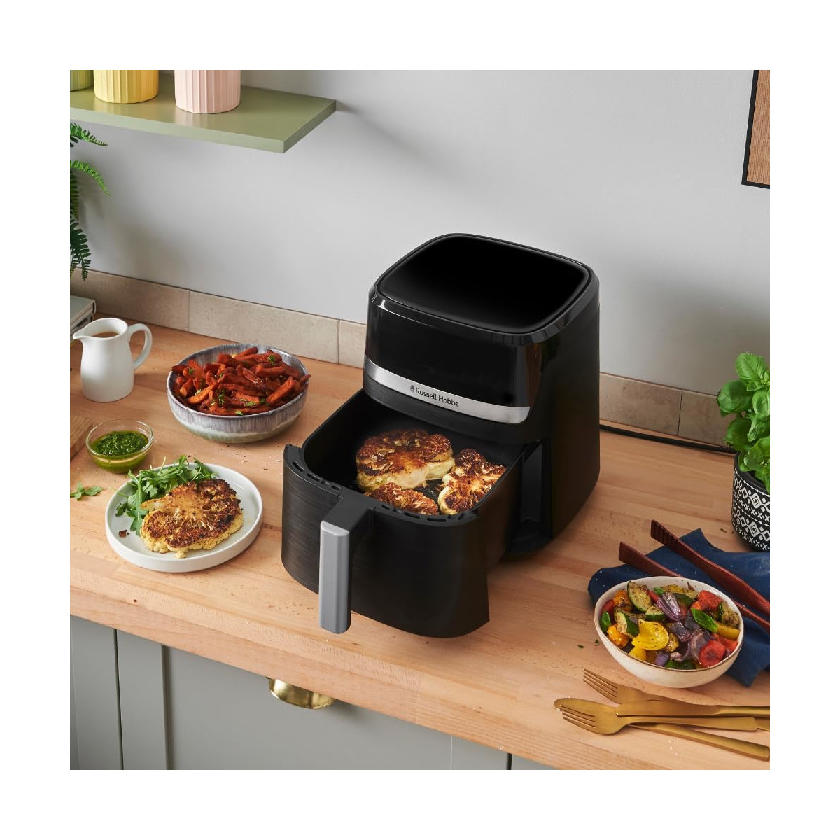 Ein schwarzer Airfryer mit Essen darin, umgeben von gekochtem Gemüse, einem Krug und einem Teller mit Essen auf einer Holztheke.