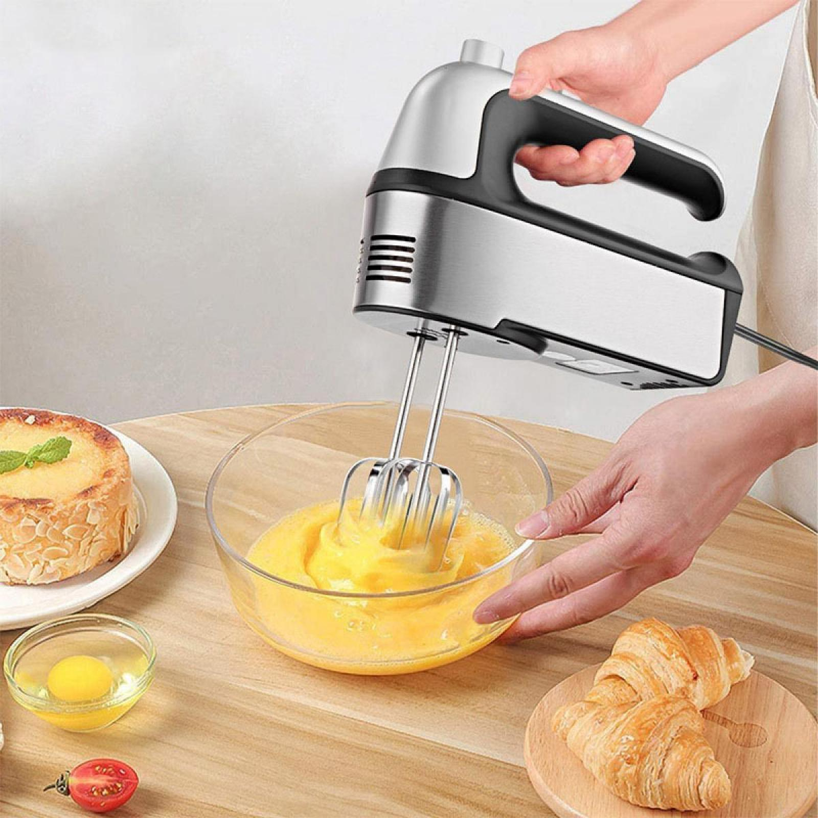 Ein Handmixer, der Eier in einer Schüssel verrührt. Croissants, eine Torte und eine Tomate liegen auch auf dem Tisch.
