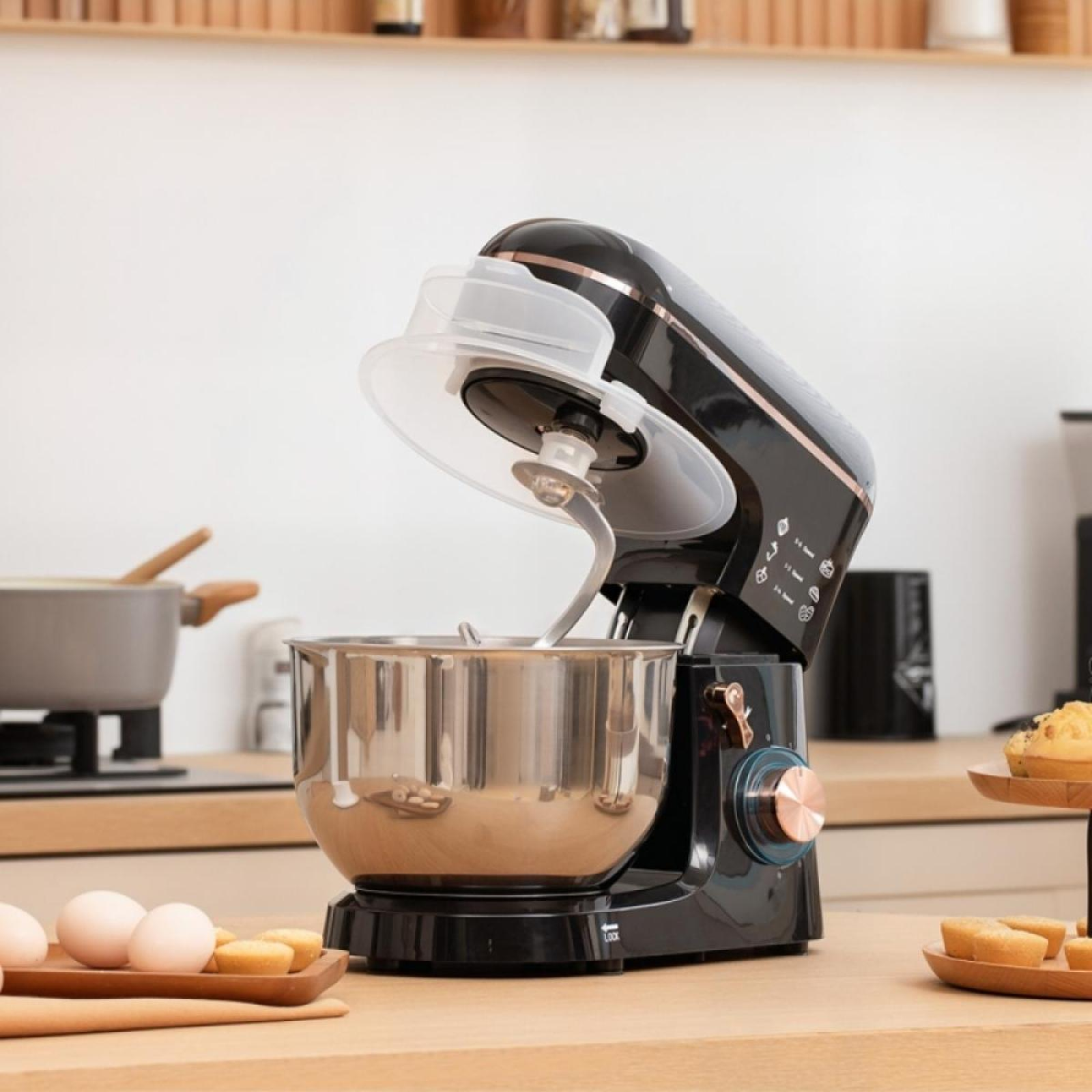 Ein schwarzer Küchenmixer mit einer Edelstahlschüssel steht auf einer Holztheke. Eier und kleine Kuchen sind ebenfalls vorhanden.