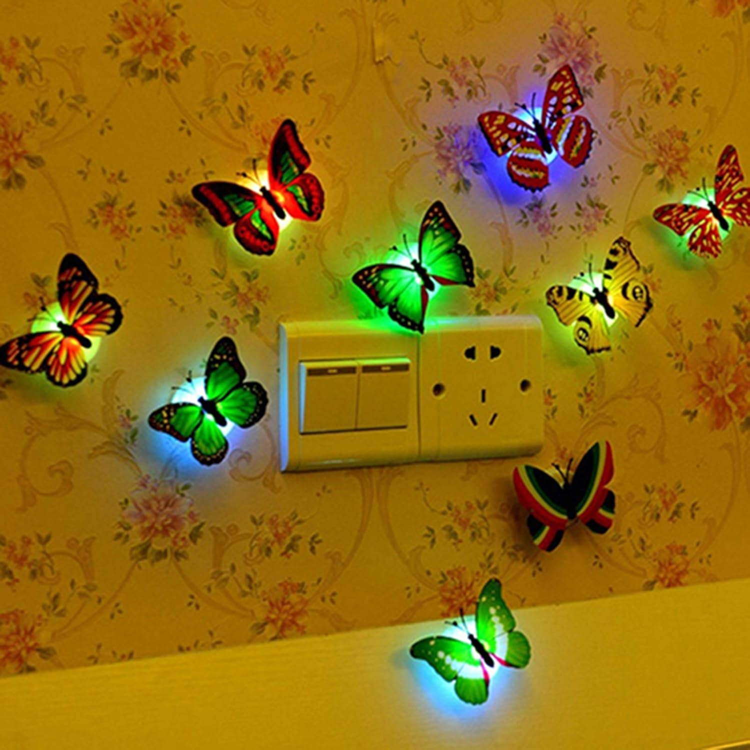 Bunte Schmetterlingslichter auf Tapete mit Blumenmuster. Ein Lichtschalter und eine Steckdose sind sichtbar. Schmetterlinge sind grün, rot, gelb und blau.