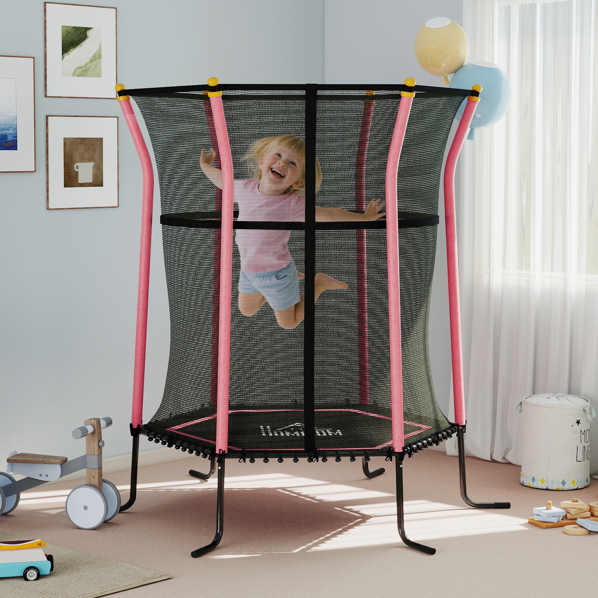 Una bambina salta su un trampolino. È rosa e nero. Ci sono dei palloncini di lato. Altri giocattoli sono sul pavimento.