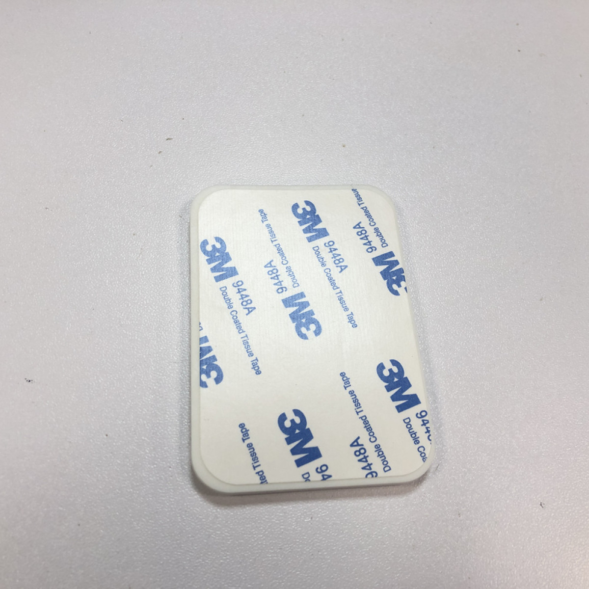Biała, prostokątna etykieta taśmy dwustronnej 3M. Etykieta ma niebieskie logo i tekst 3M. Znajduje się na jasnoszarej powierzchni.