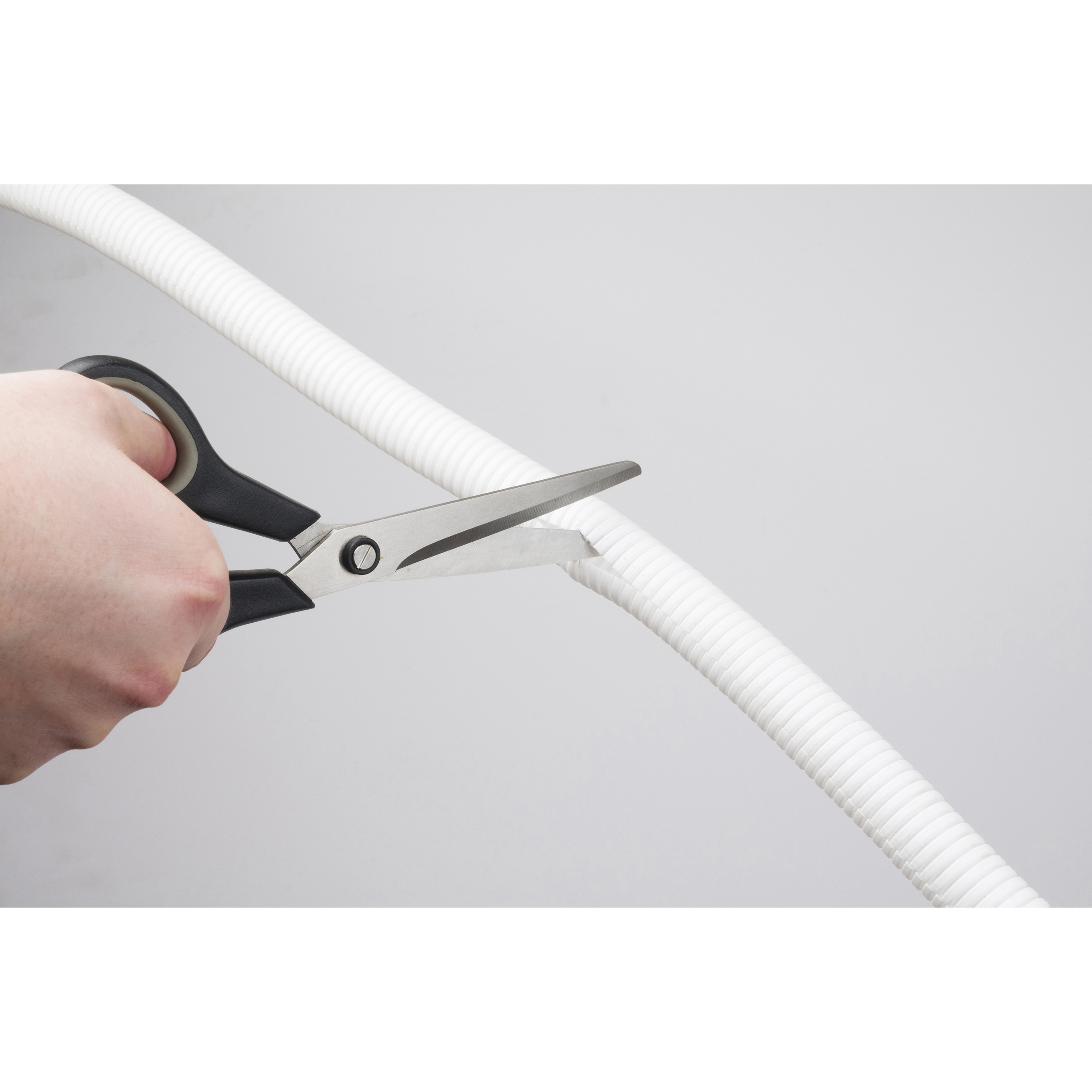 Mano con forbici che taglia un tubo di plastica ondulato bianco su sfondo grigio.