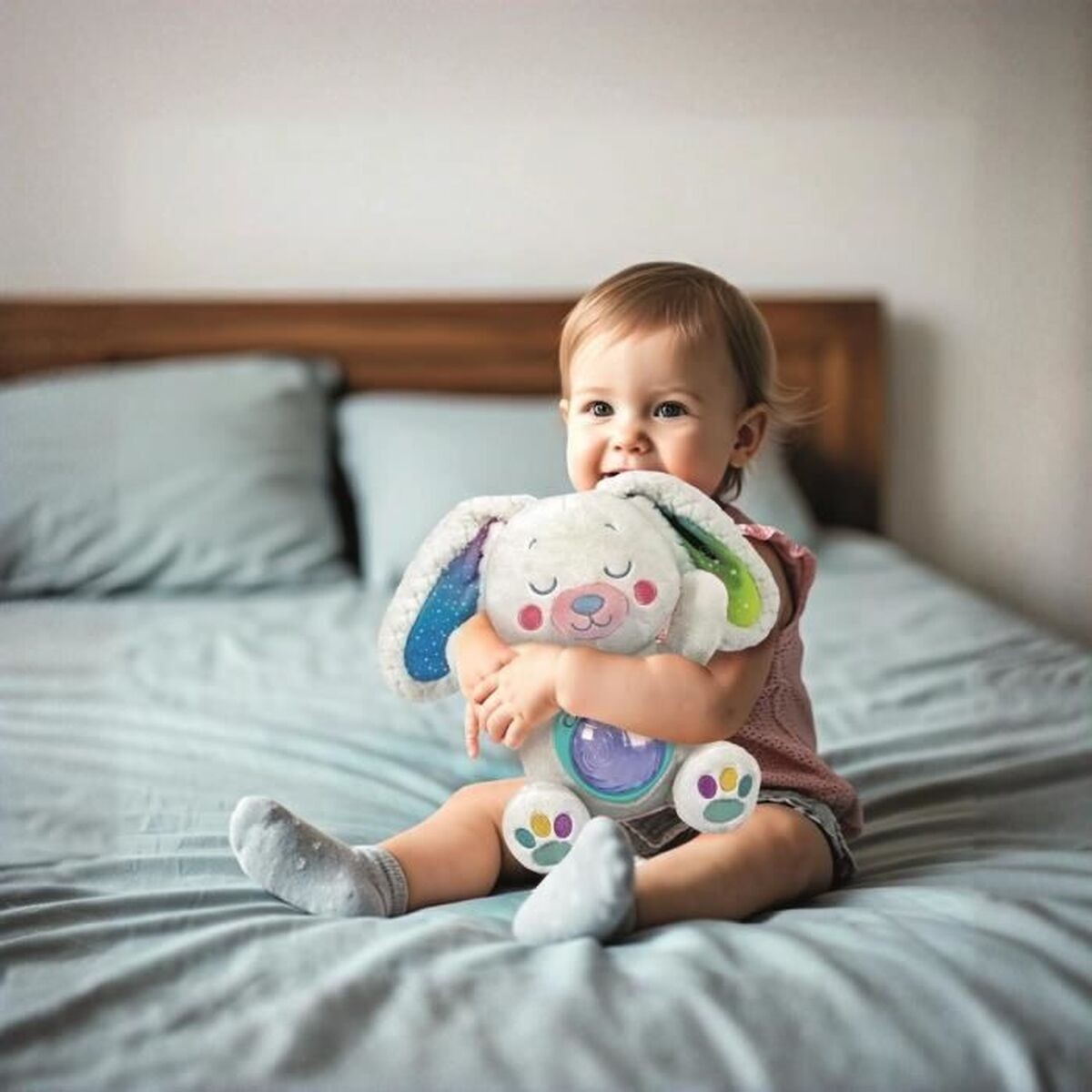 Un bambino seduto su un letto abbraccia un peluche a forma di coniglietto bianco. Il bambino sorride. Il letto ha lenzuola e cuscini blu.