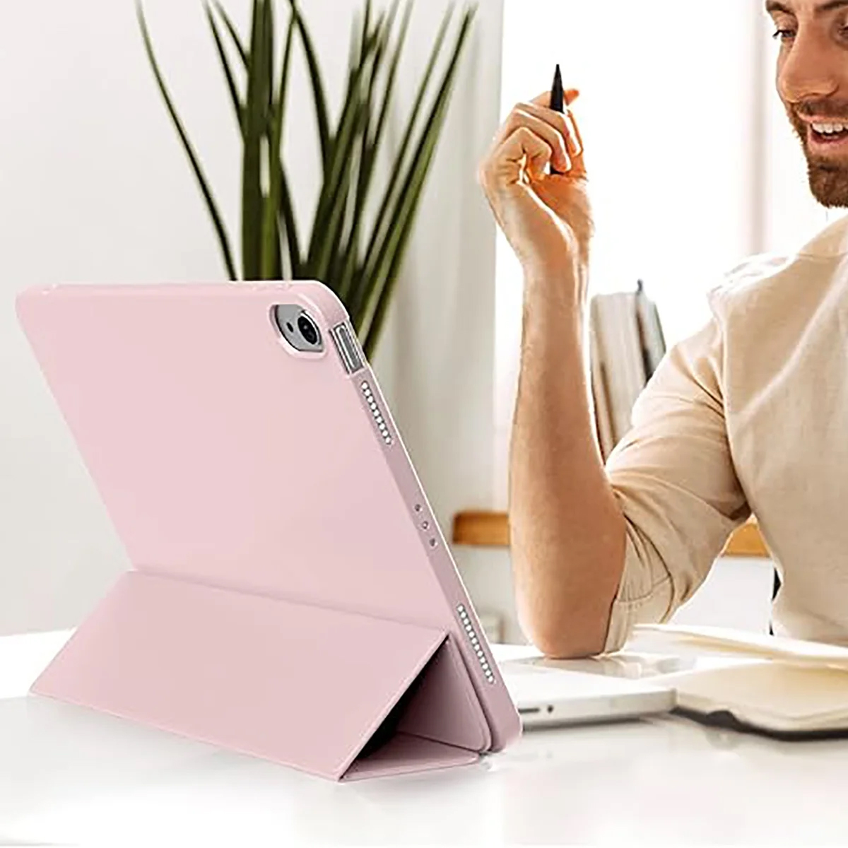 Ein rosa Tablet steht auf einem weißen Schreibtisch. Ein Mann hält einen Stift und lächelt. Grüne Pflanzen sind im Hintergrund.