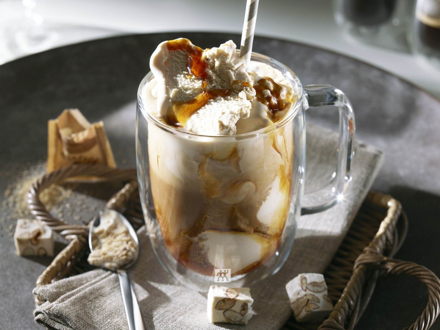 Kaffeegetränk im Glasbecher mit Eis und Strohhalm. Nougat, Löffel und Serviette auf einem Tablett. Karamellregen oben.