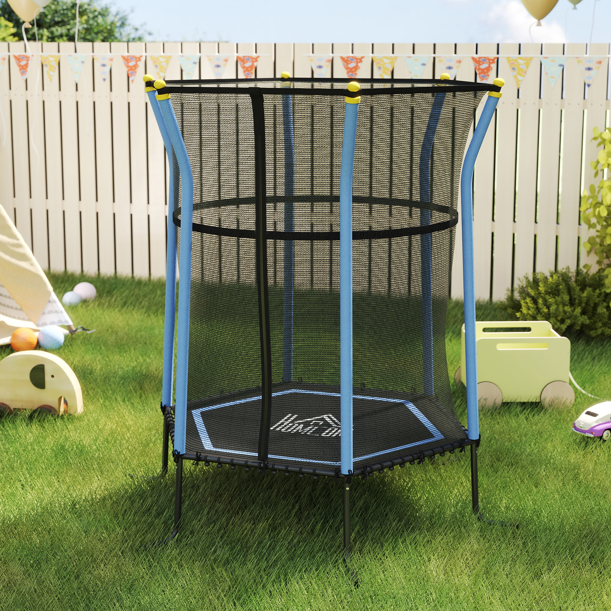 Un trampolino blu in un cortile erboso con una staccionata bianca, giocattoli, striscione per feste e palloncini.