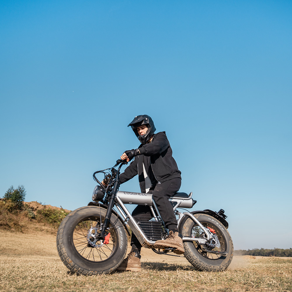 Osoba w kasku siedzi na srebrnym motocyklu na ziemi. Niebo jest niebieskie i czyste. Motocykl jest w ruchu.