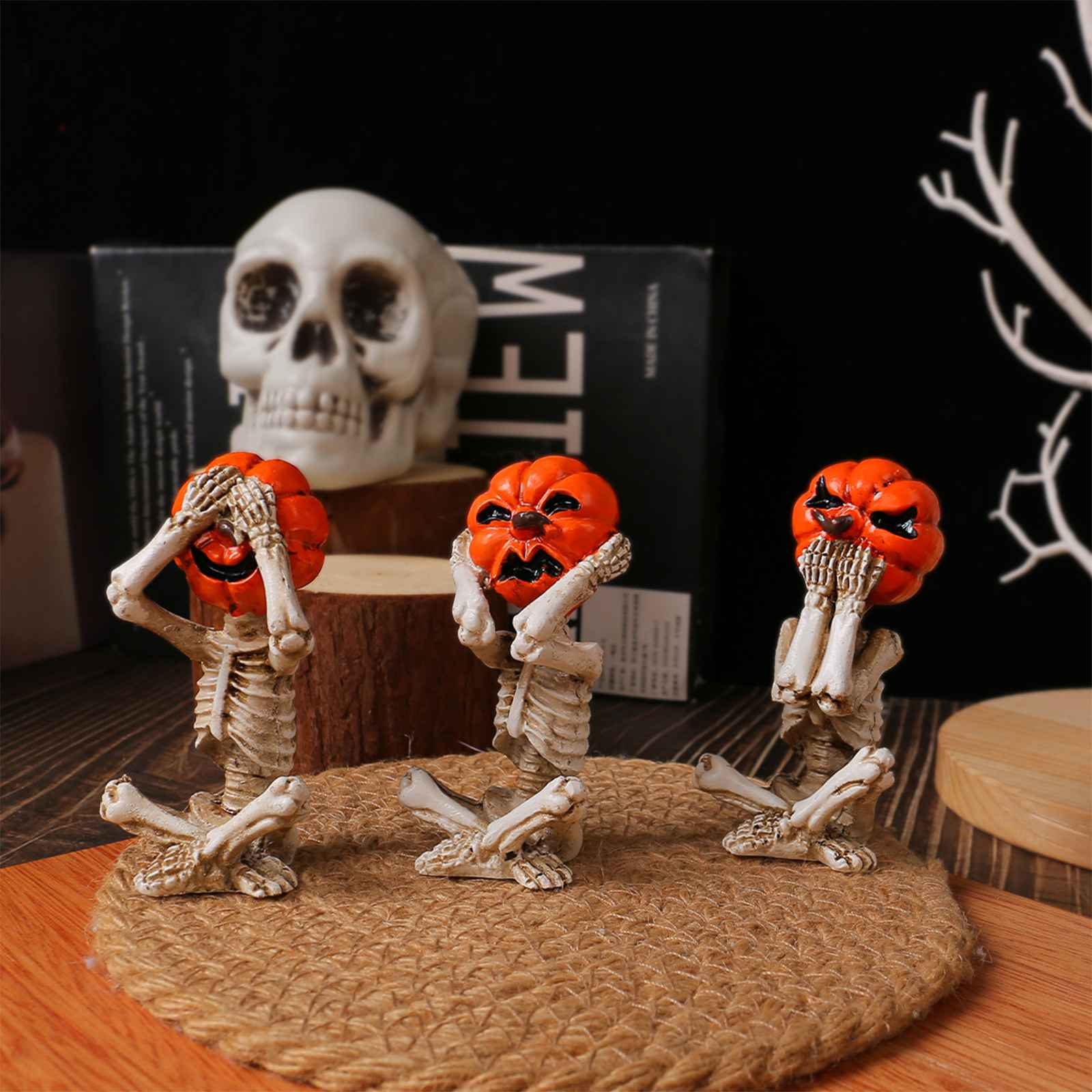 Drei Skelettfiguren mit Kürbisköpfen, die sich die Augen und Münder bedecken. Ein Totenkopf und Bücher sind im Hintergrund.