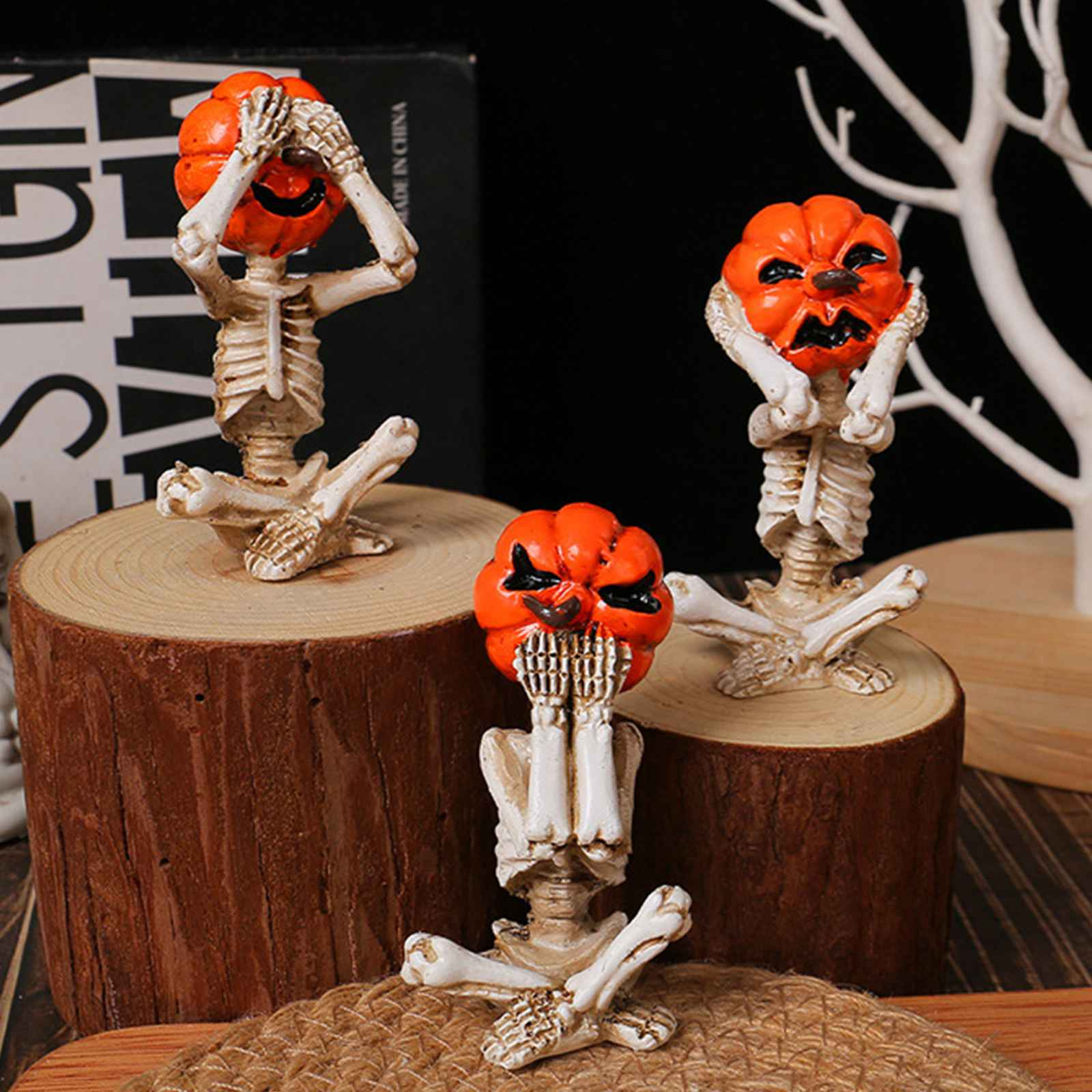 Drei Skelettfiguren mit Kürbisköpfen sitzen auf Holzscheiben. Im Hintergrund ein Buch und ein weißer Baum.