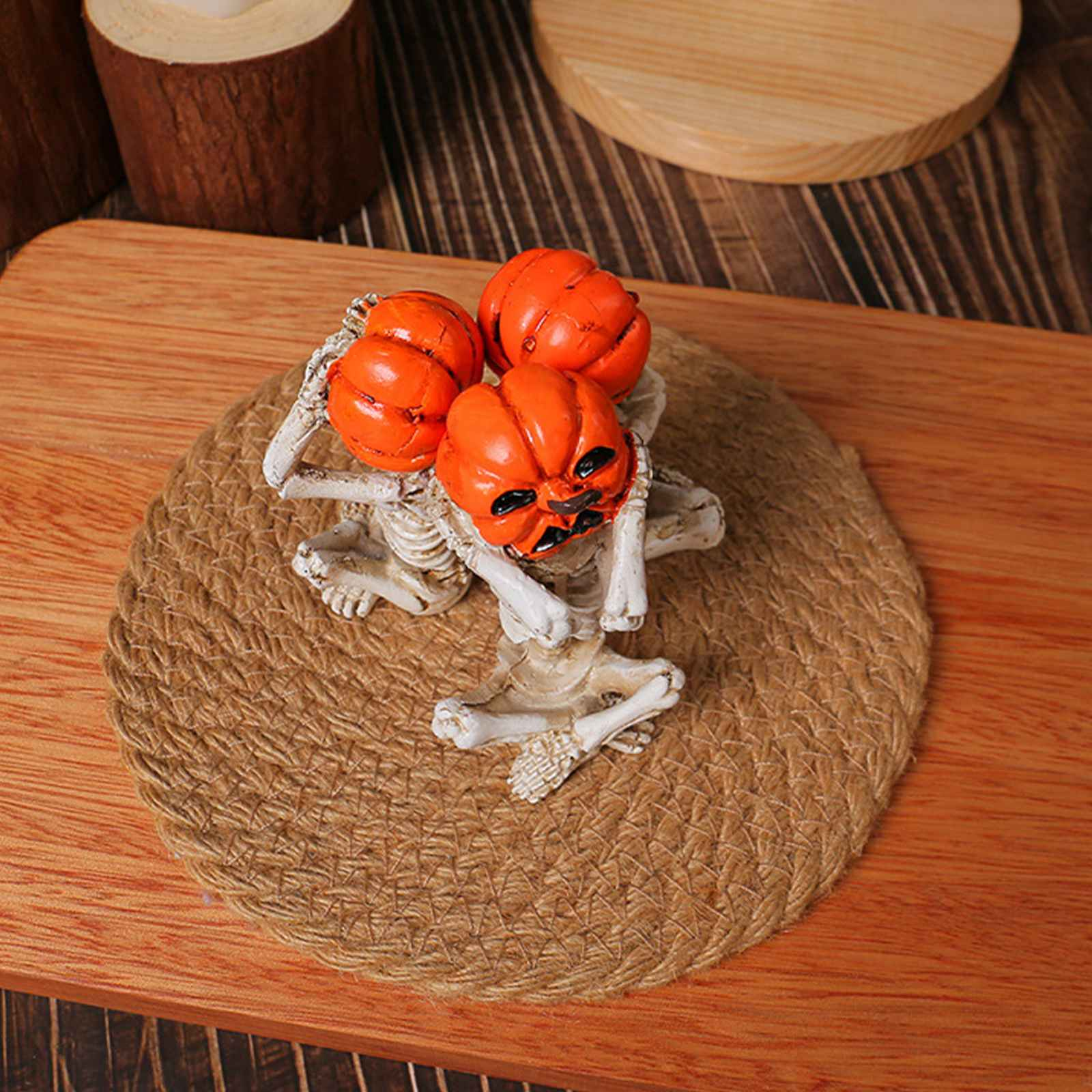Eine Skelettfigur hält drei orangefarbene Kürbisse auf einem Jute-Untersetzer auf einer Holzoberfläche.