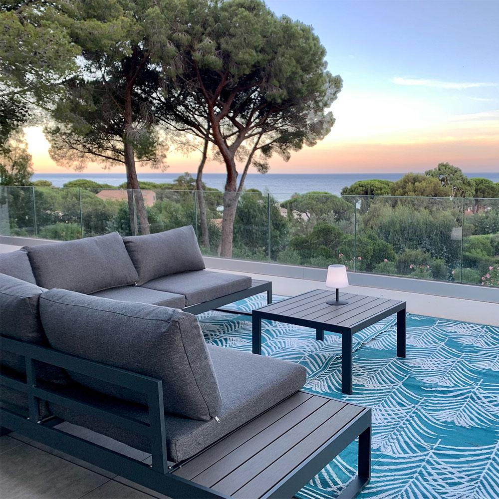 Terrasse mit grauen Möbeln, Tisch und Teppich mit Blättermuster. Bäume und Meer im Hintergrund. Sonnenuntergangshimmel.