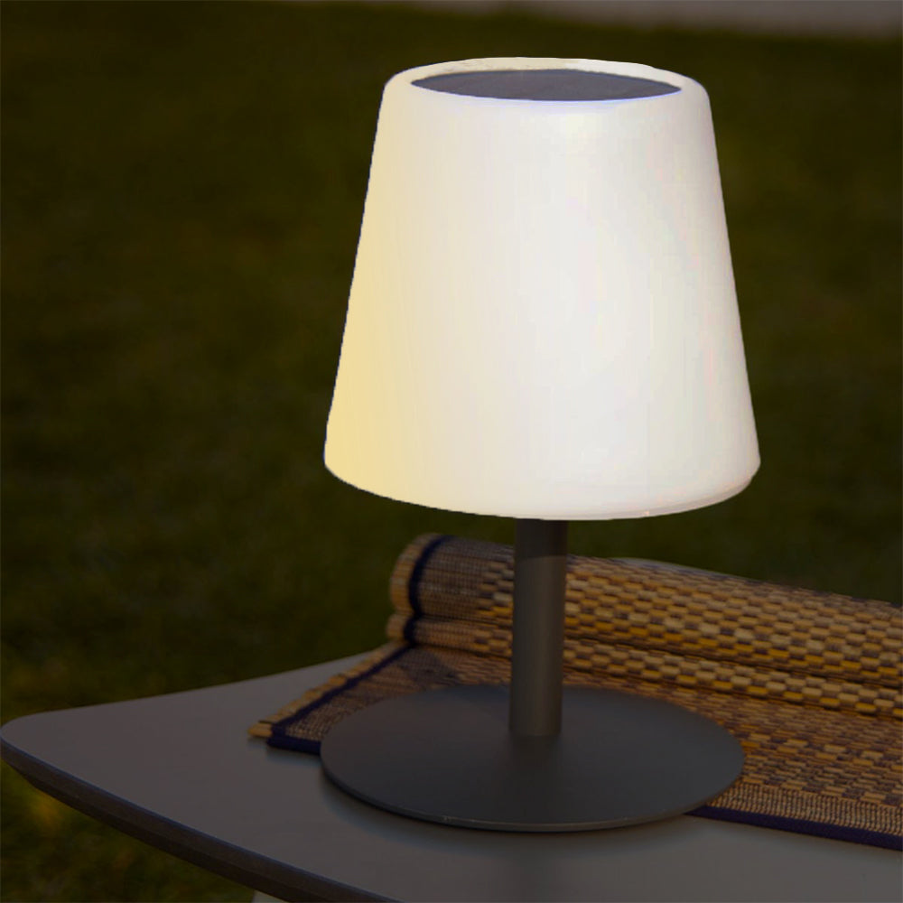 Eine weiße Lampe mit schwarzem Sockel und dunkelgrünem Hintergrund, die auf einem grauen Tisch steht. Die Lampe steht auf einer gewebten Matte.