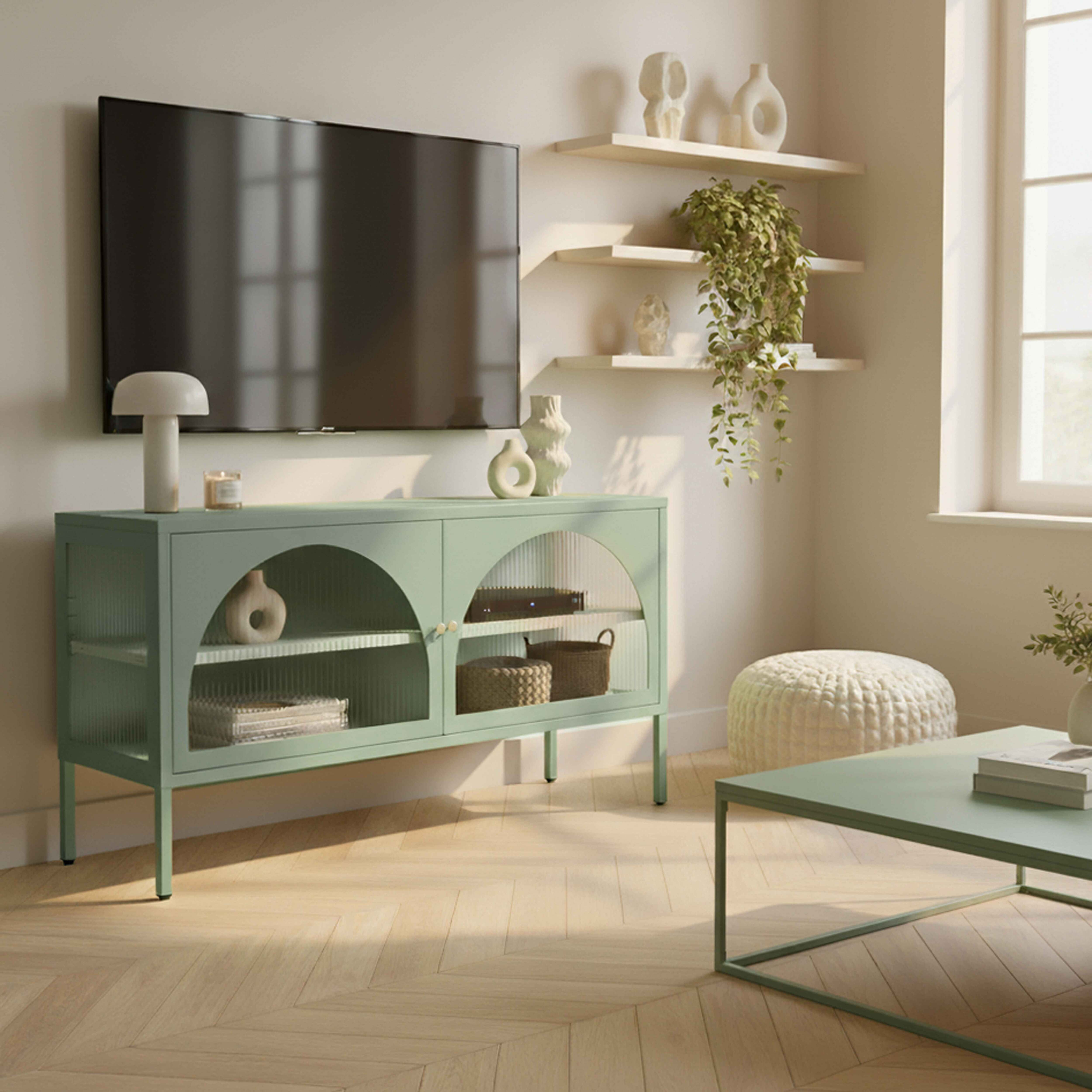 Ein Wohnzimmer mit Fernseher, grünem Schrank, Regalen mit Dekoration und einem Couchtisch. Holzboden und Fenster.