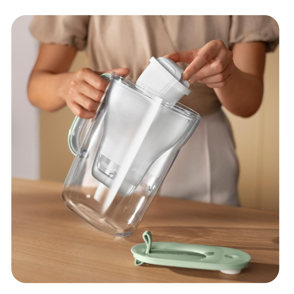 Eine Person setzt einen weißen Wasserfilter in einen durchsichtigen Krug ein. Ein grüner Deckel und ein Holztisch sind im Vordergrund.