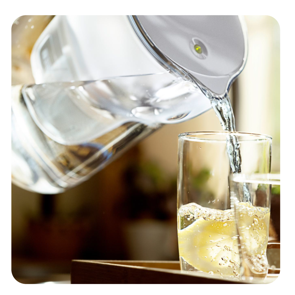 Wasser gießt aus einem Krug in ein Glas mit einer Zitronenscheibe. Blasen bilden sich. Unter dem Glas ist ein Holztablett zu sehen.
