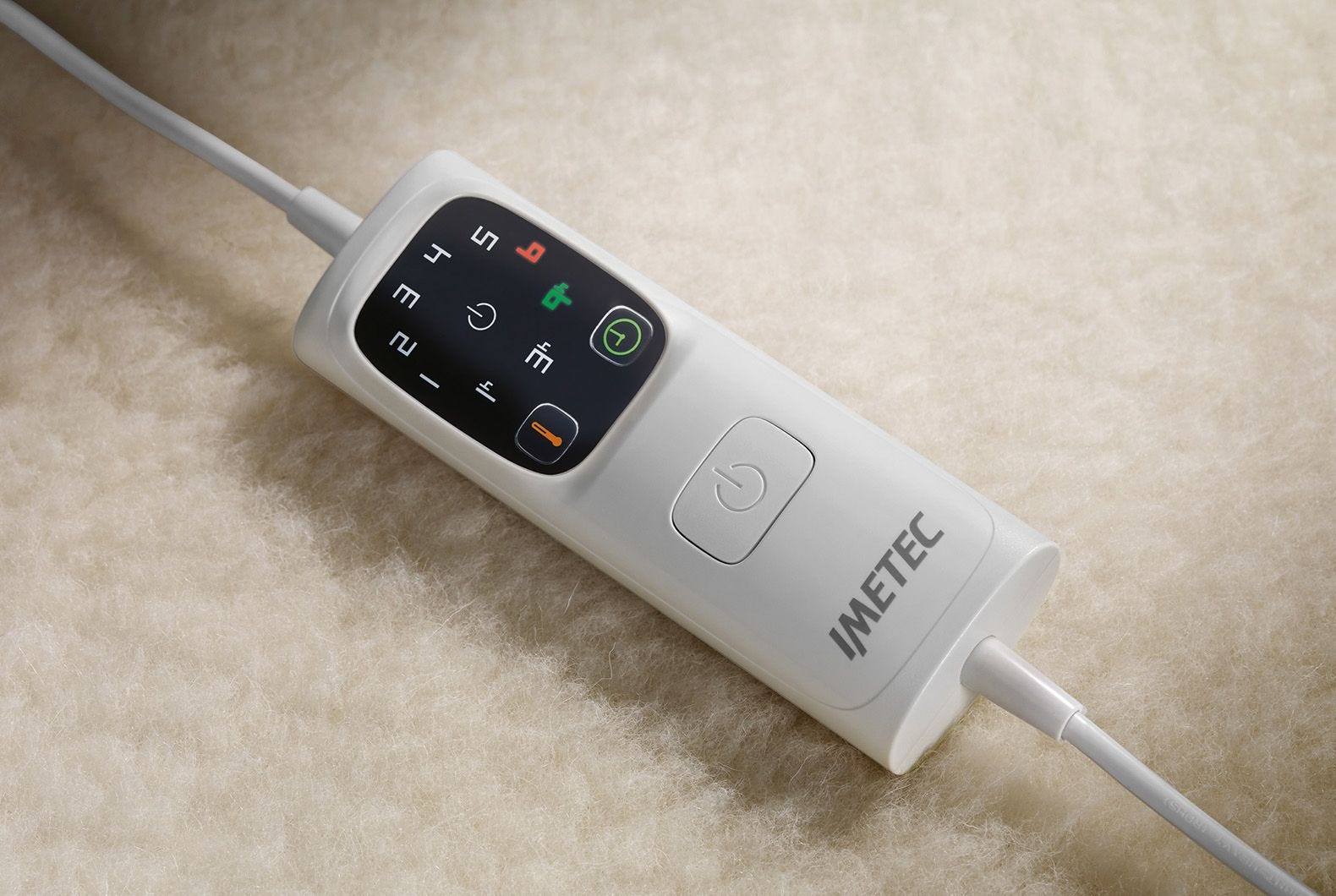 Un panel de control blanco con botones y pantalla, conectado a un cable blanco, descansa sobre una superficie beige y esponjosa.