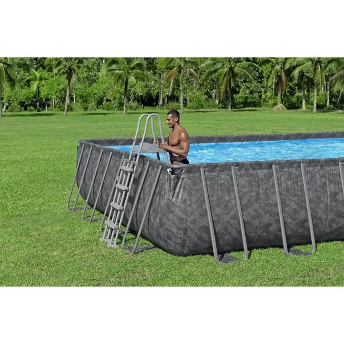 Un uomo è in una piscina, tenendo la scala. La piscina è in un giardino verde con palme sullo sfondo.