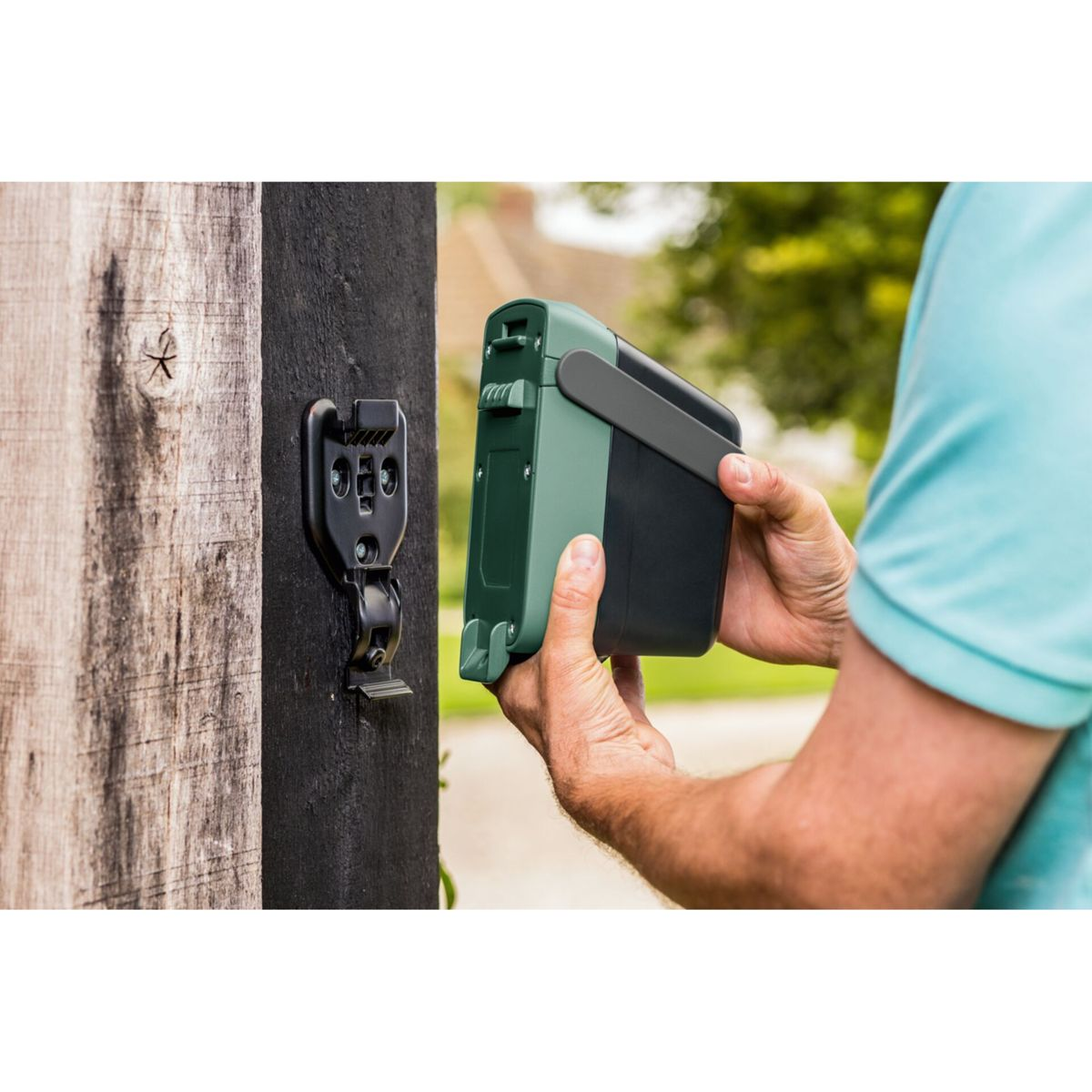 Osoba trzymająca zielono-czarne urządzenie w pobliżu drewnianego słupka z przymocowanym czarnym wspornikiem. Tło przedstawia zieleń i dom.