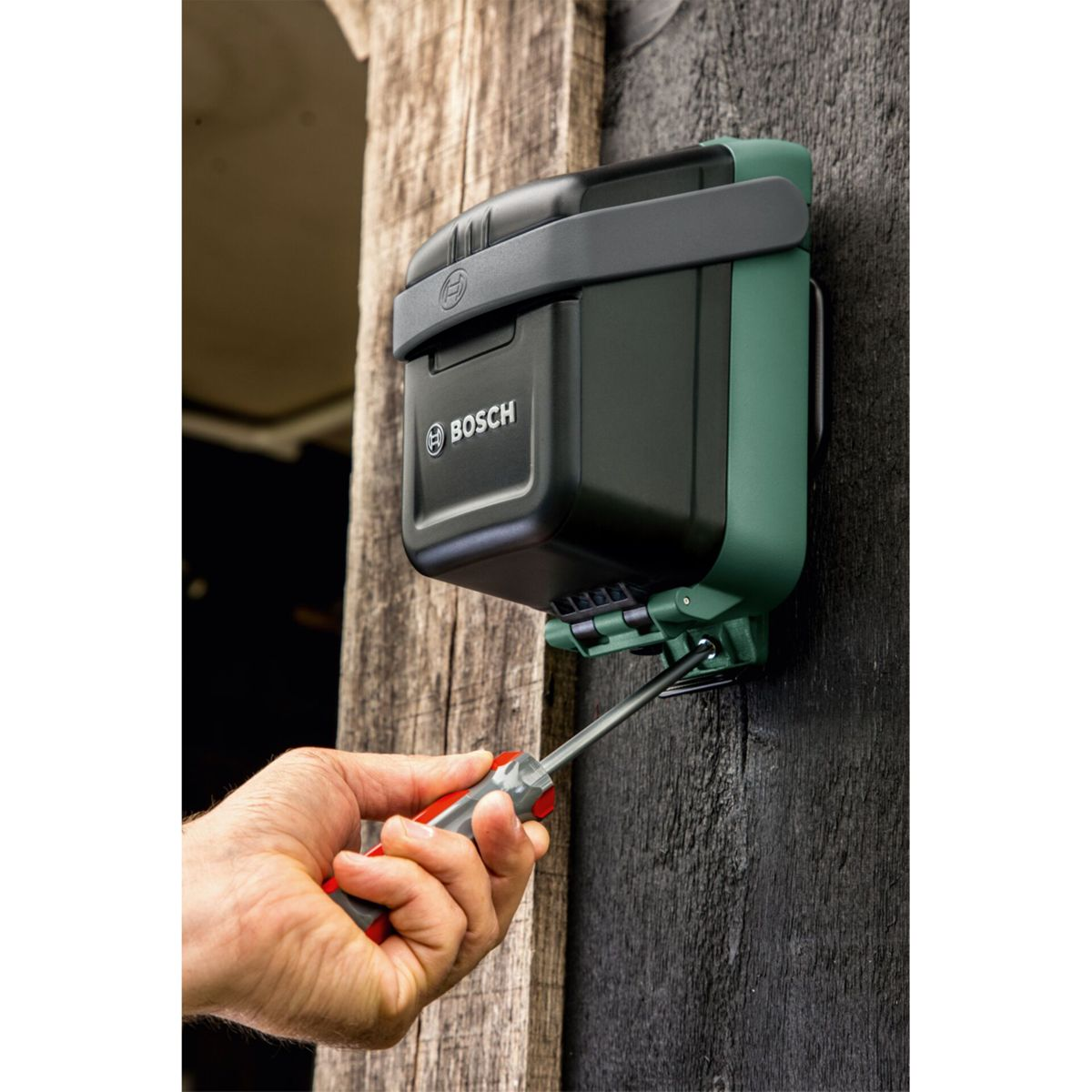Ręka używa śrubokręta do zamocowania zielono-czarnego urządzenia Bosch na drewnianej ścianie.