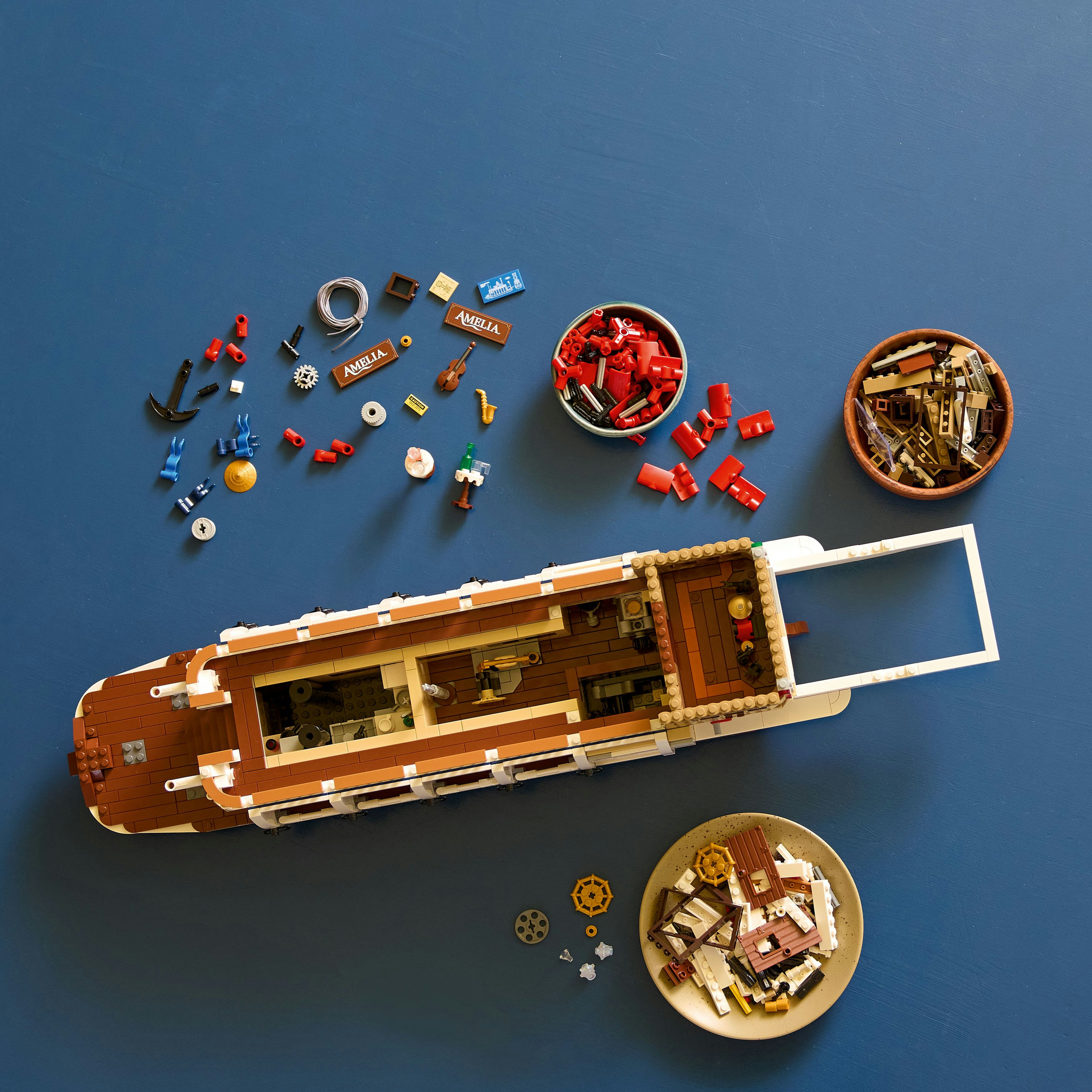 Łódź Lego częściowo złożona, z rozrzuconymi elementami i miskami klocków na niebieskiej powierzchni. Łódź jest brązowa i biała.